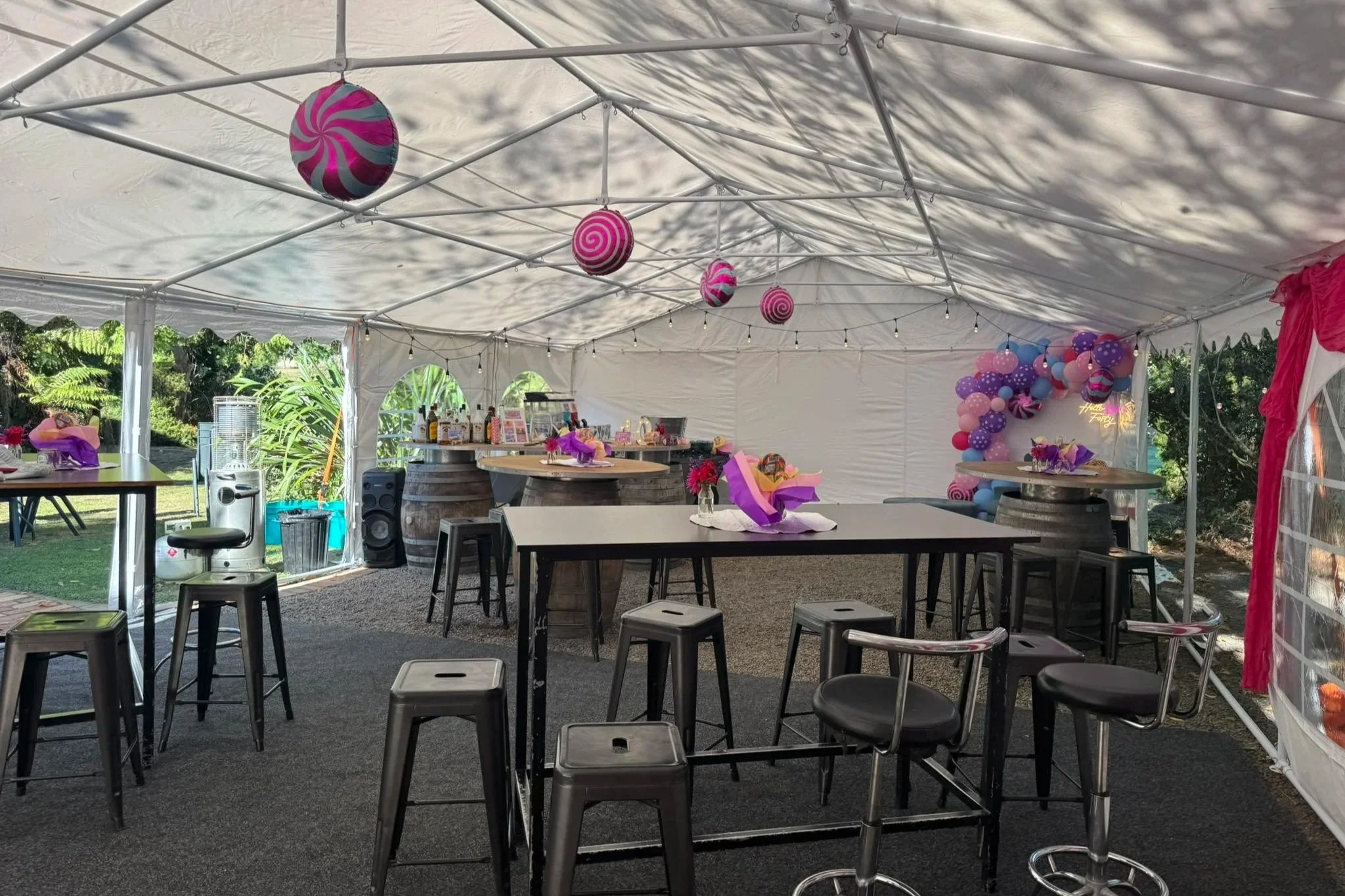 Indoor party tent decorated with pink and purple balloons, hanging lanterns, and tables with flowers and gifts, outdoor greenery visible through open sides