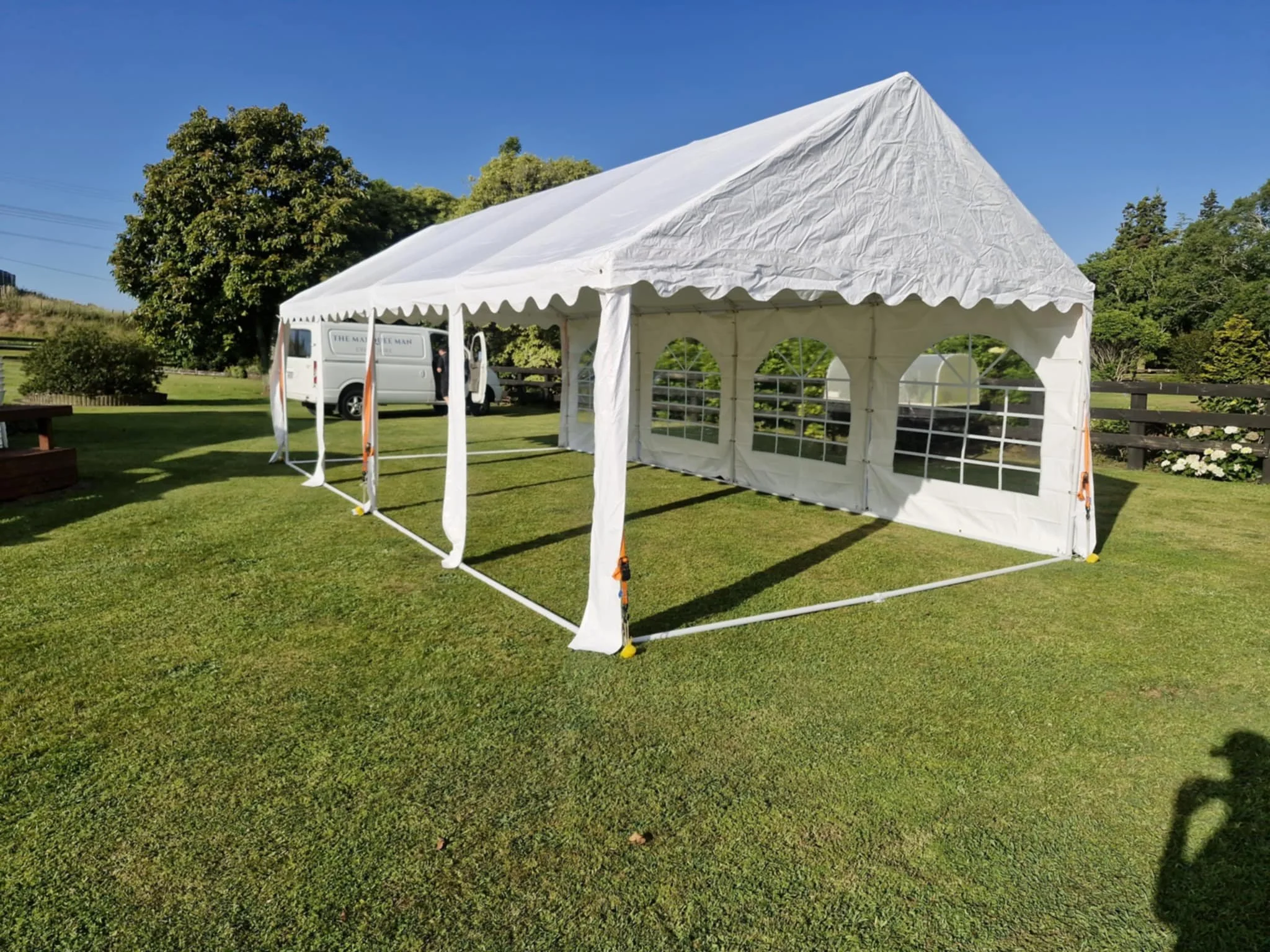 A large white outdoor event tent with transparent windows on a grassy area, set up on a sunny day with trees and blue sky in the background.