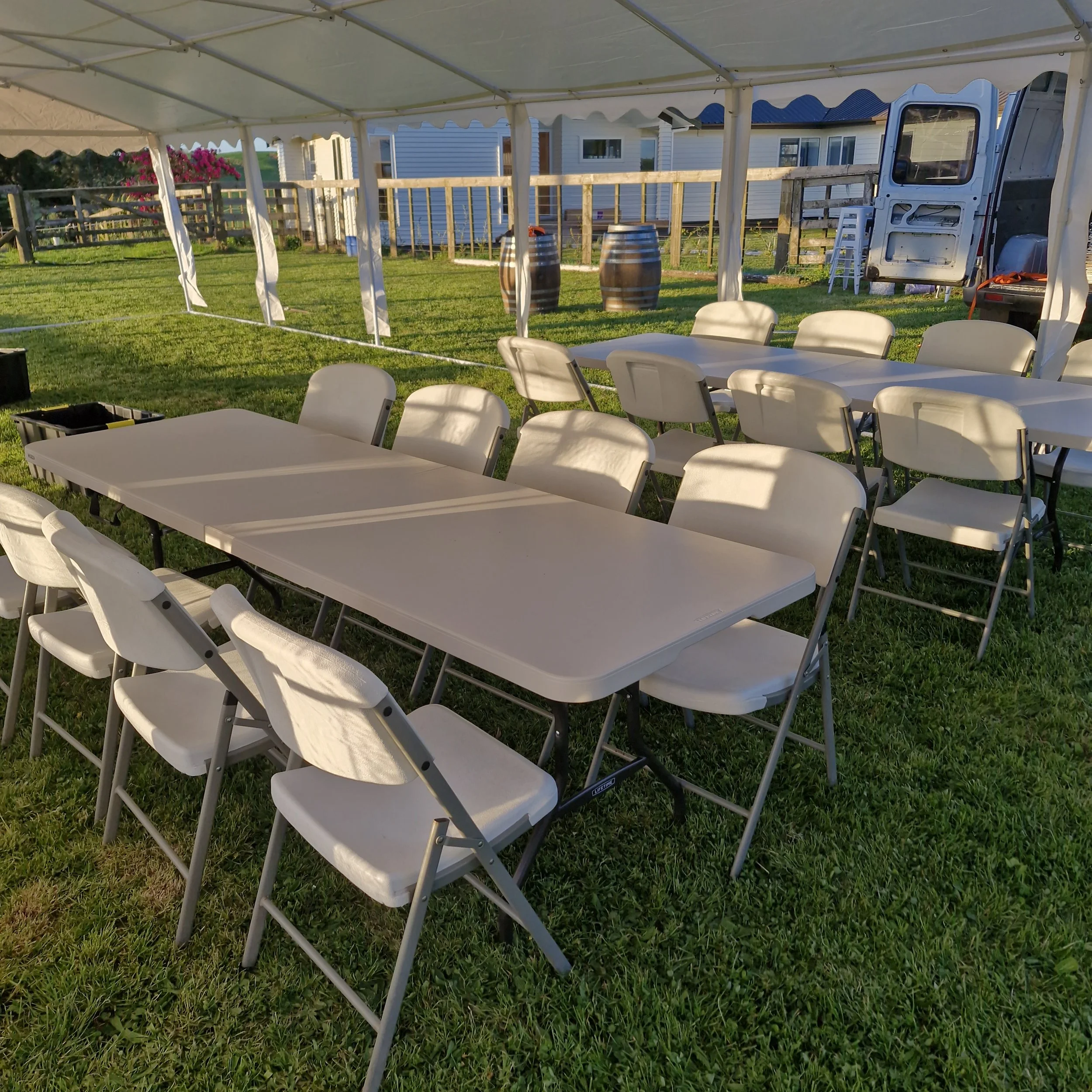 Outdoor event setup with white folding chairs around white tables inside a large tent, with barrels, a fence, a house in the background, and a lawn.