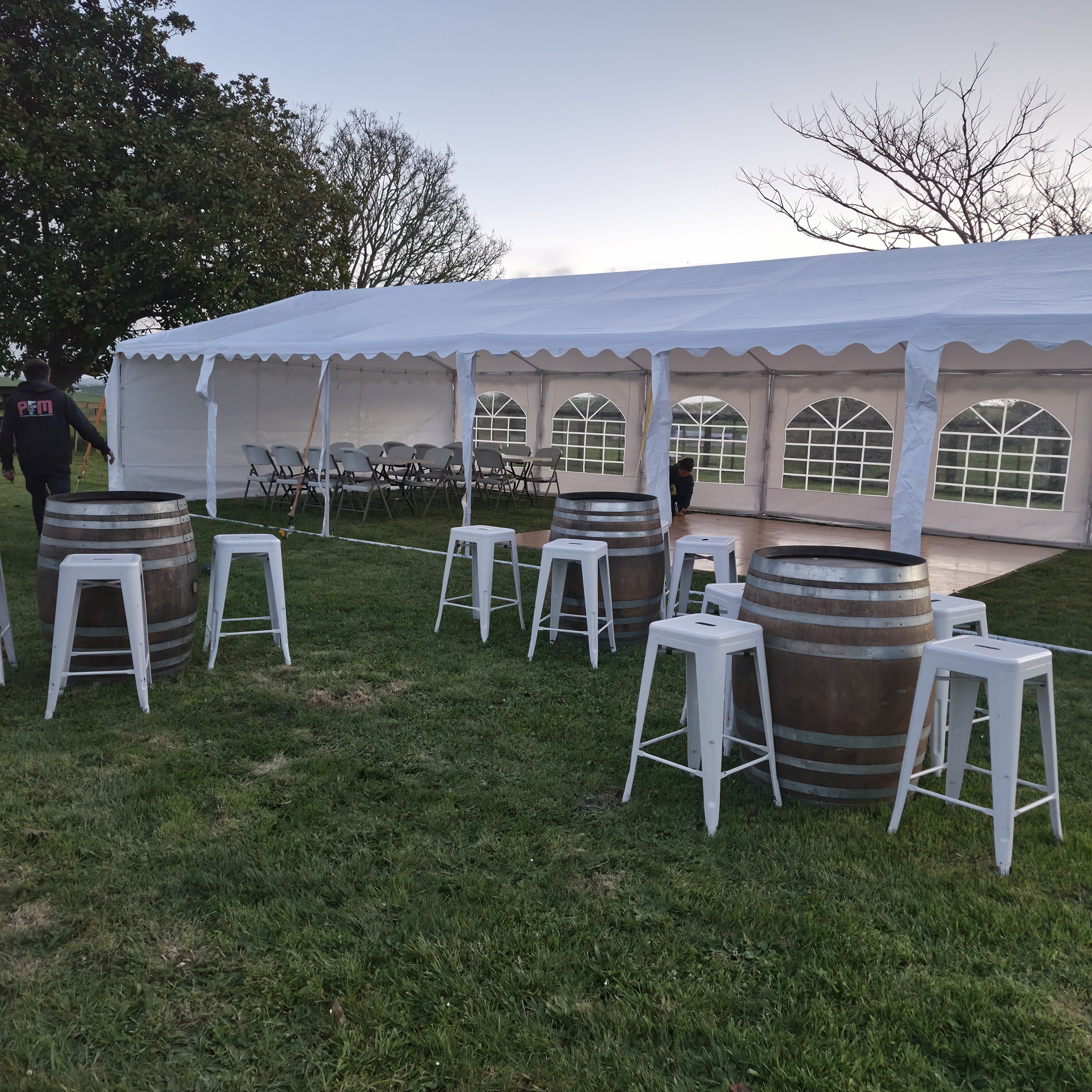A large white event tent set up outdoors on green grass, with folding chairs arranged inside. In the foreground, there are decorative wine barrel tables, white stools, and a person walking past the tent. The background includes trees with bare branch
