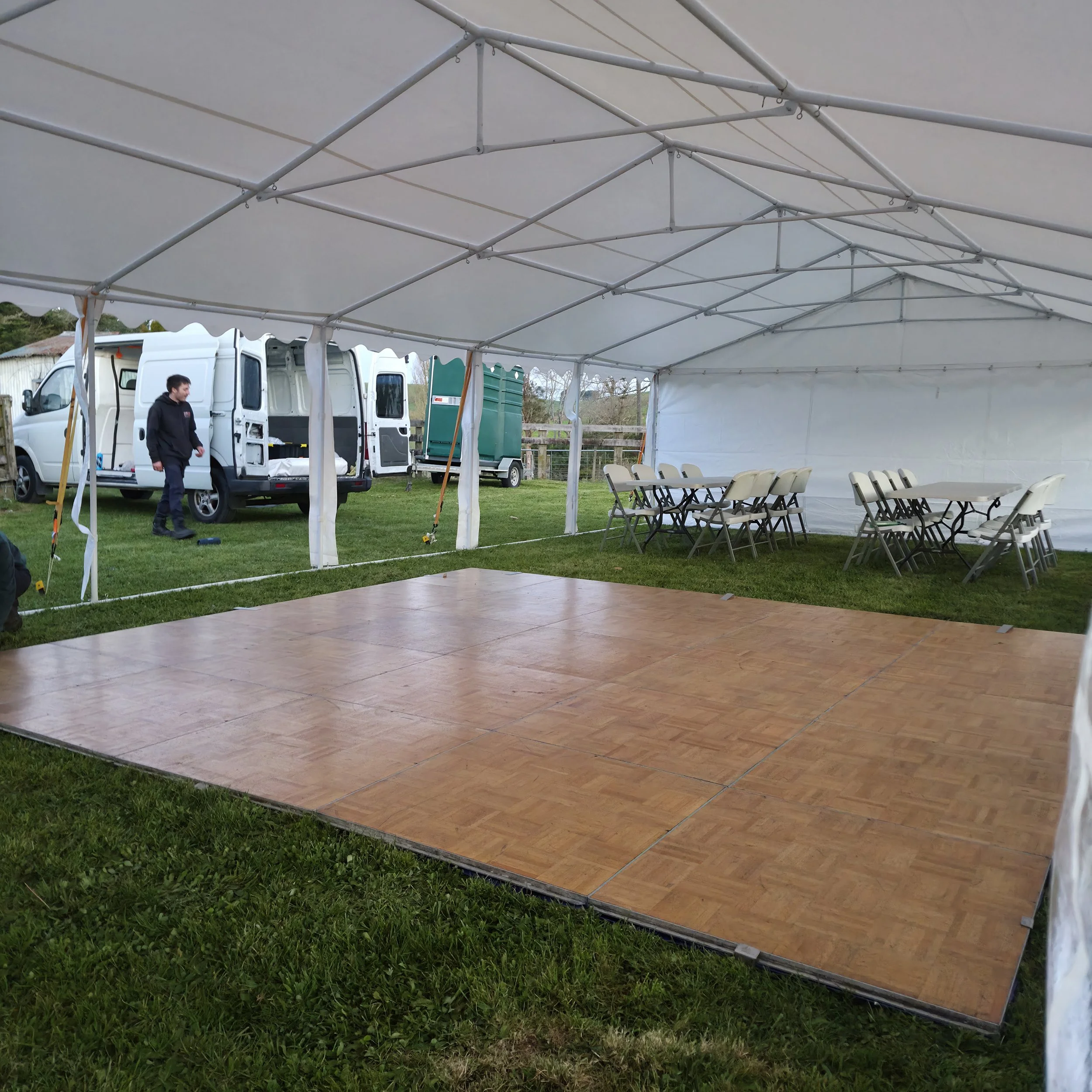 An outdoor event tent with white fabric and metal support structure, set up on a grassy area. Inside the tent, there are rows of chairs and a table, with a person standing outside near a white van.