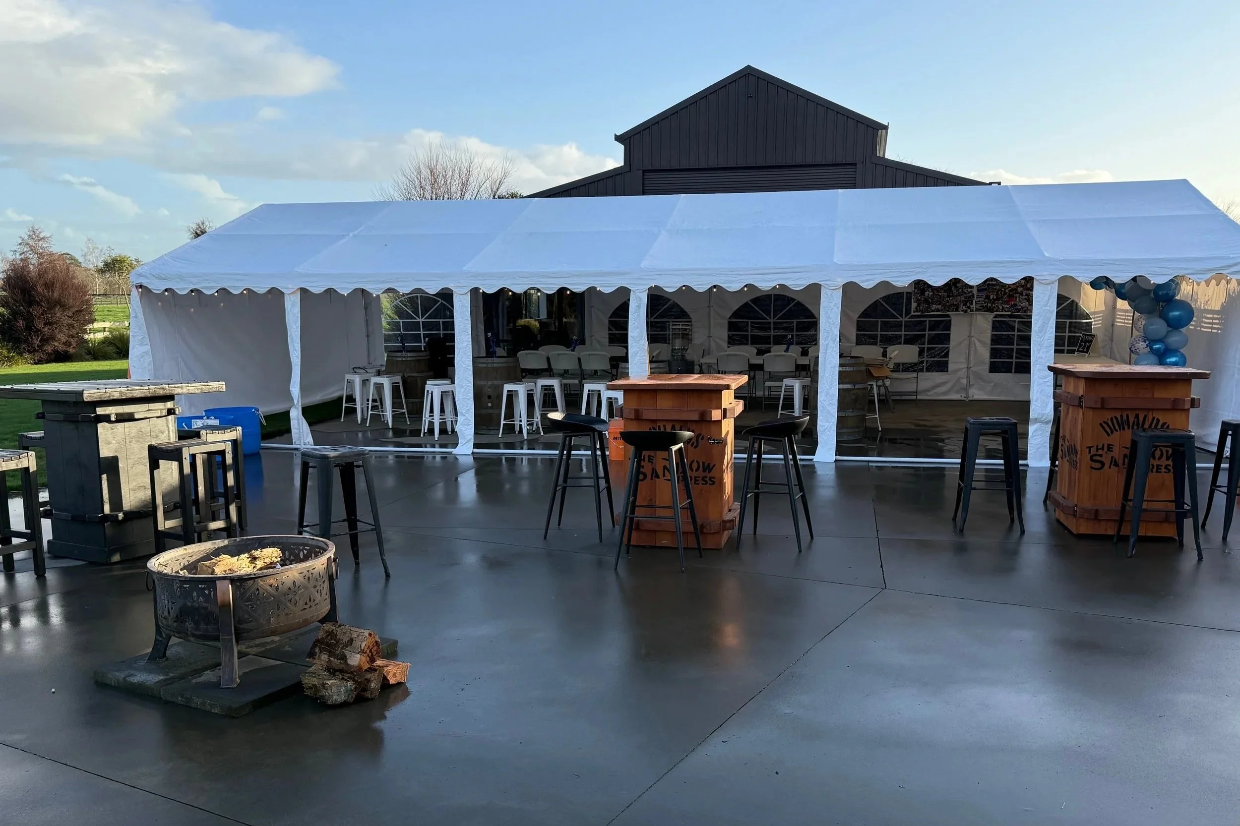 Outdoor event set up with a large white tent, high-top tables, and balloons on the right side; concrete floor with a fire pit in the foreground; green lawn and trees in the background.
