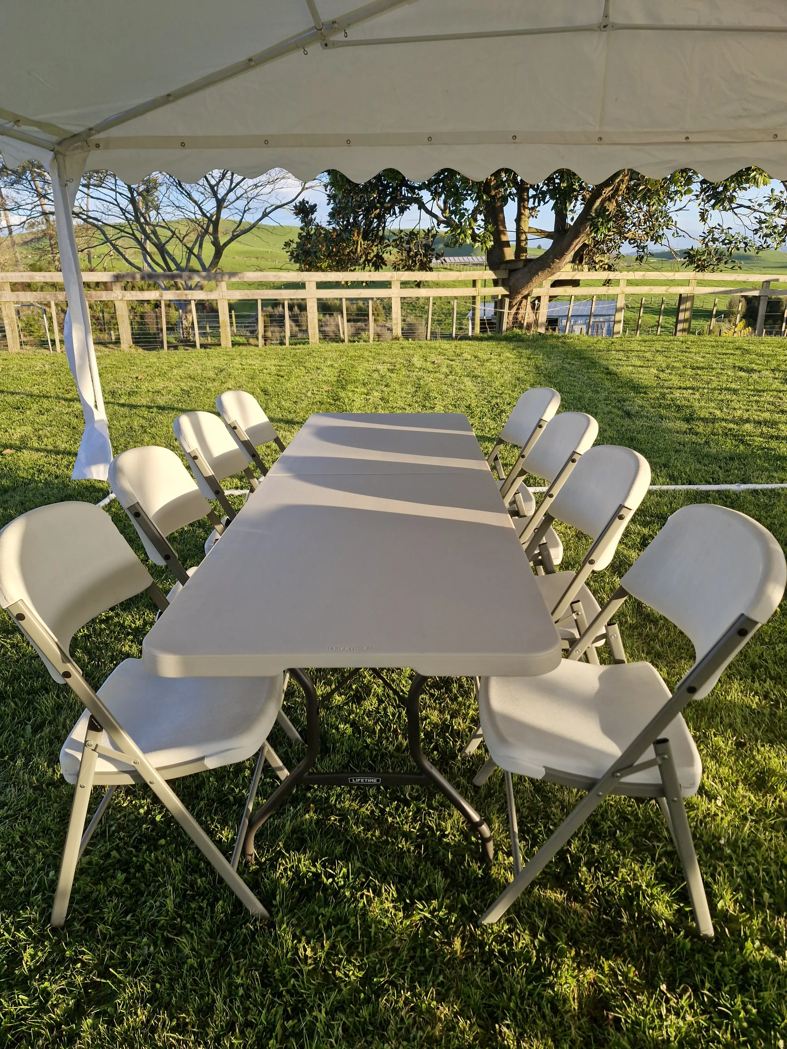An outdoor dining setup with a rectangular white table surrounded by white folding chairs on a grassy lawn. In the background, there is a wooden fence, a large tree, and rolling green hills under a clear sky.