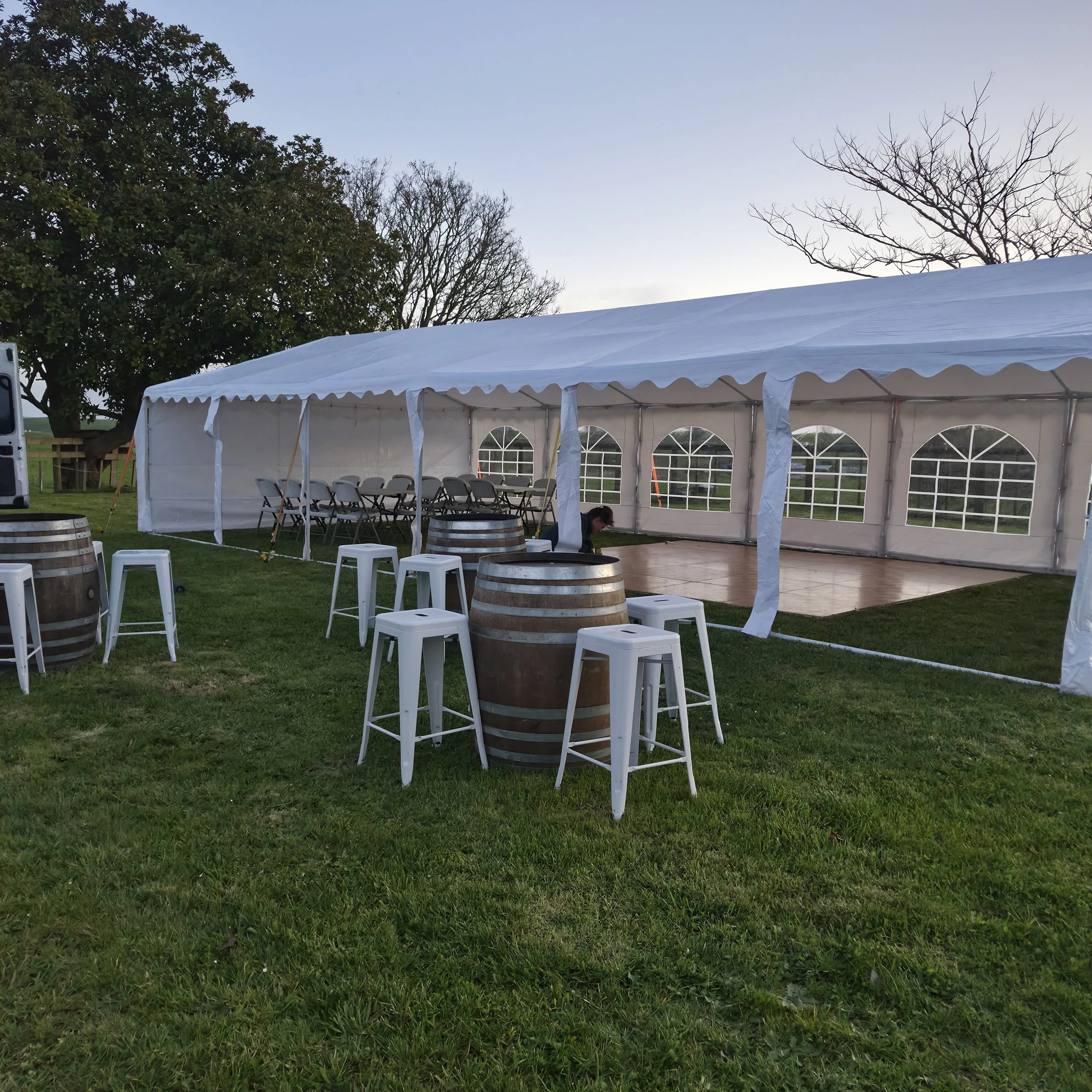 An outdoor event setup with a large white canopy tent, several chairs inside, barrels, and small white stools on a grassy area. Trees are visible in the background, and the sky is clear.