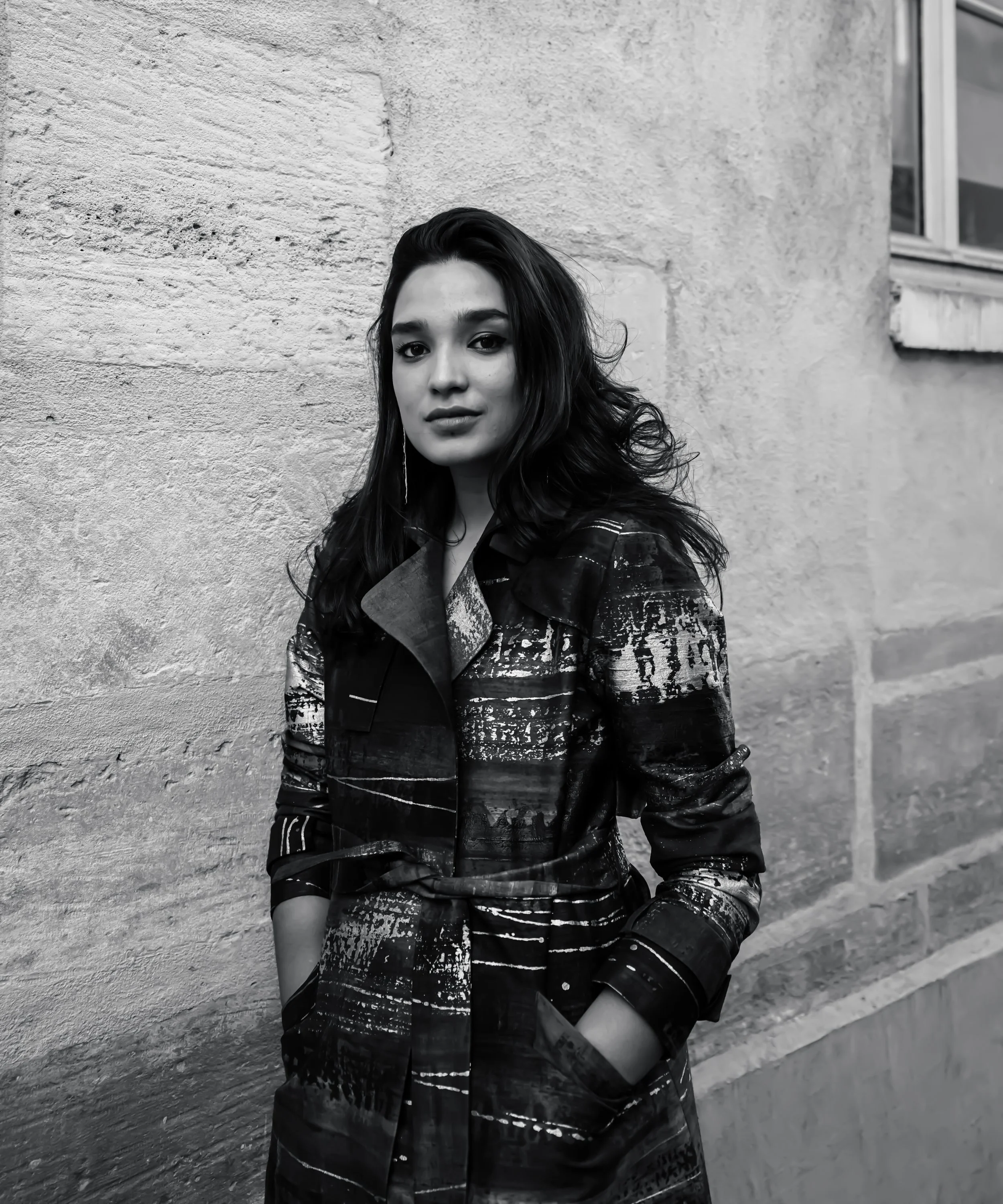 Black and white photo of a young woman standing outdoors against a textured wall with her hands in her coat pockets, looking at the camera.