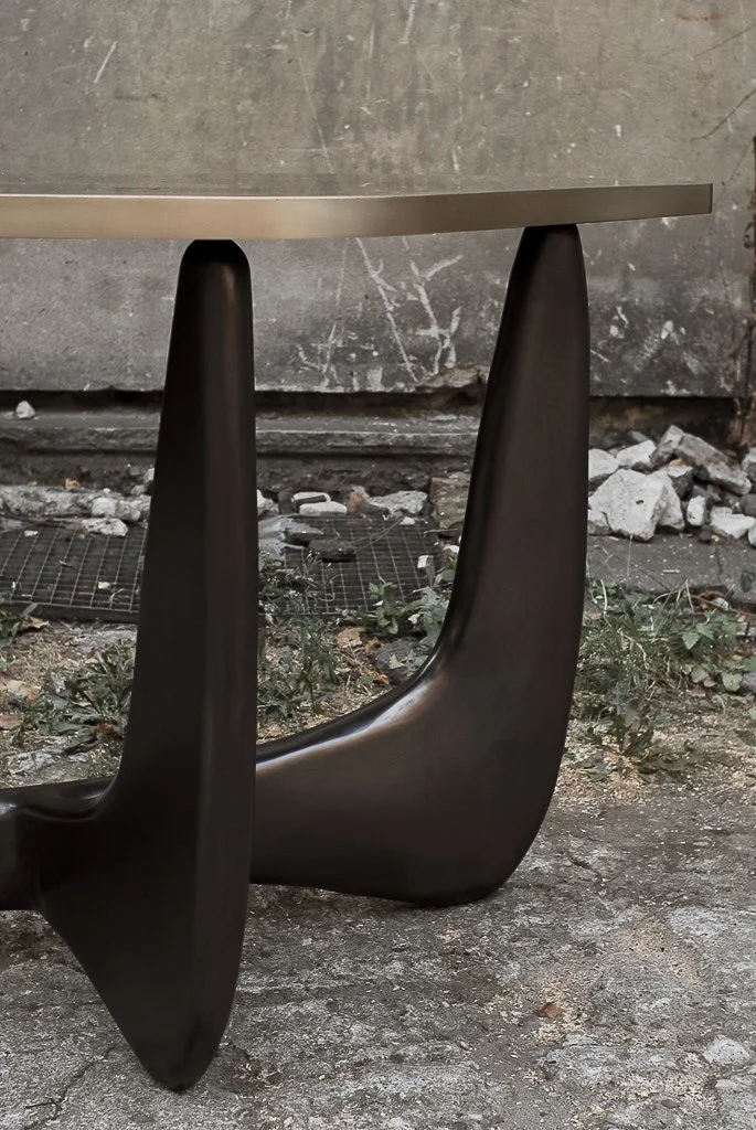 Close-up of a modern black table leg with a curved design, on an outdoor ground with dirt, small plants, and rubble, against a concrete wall.