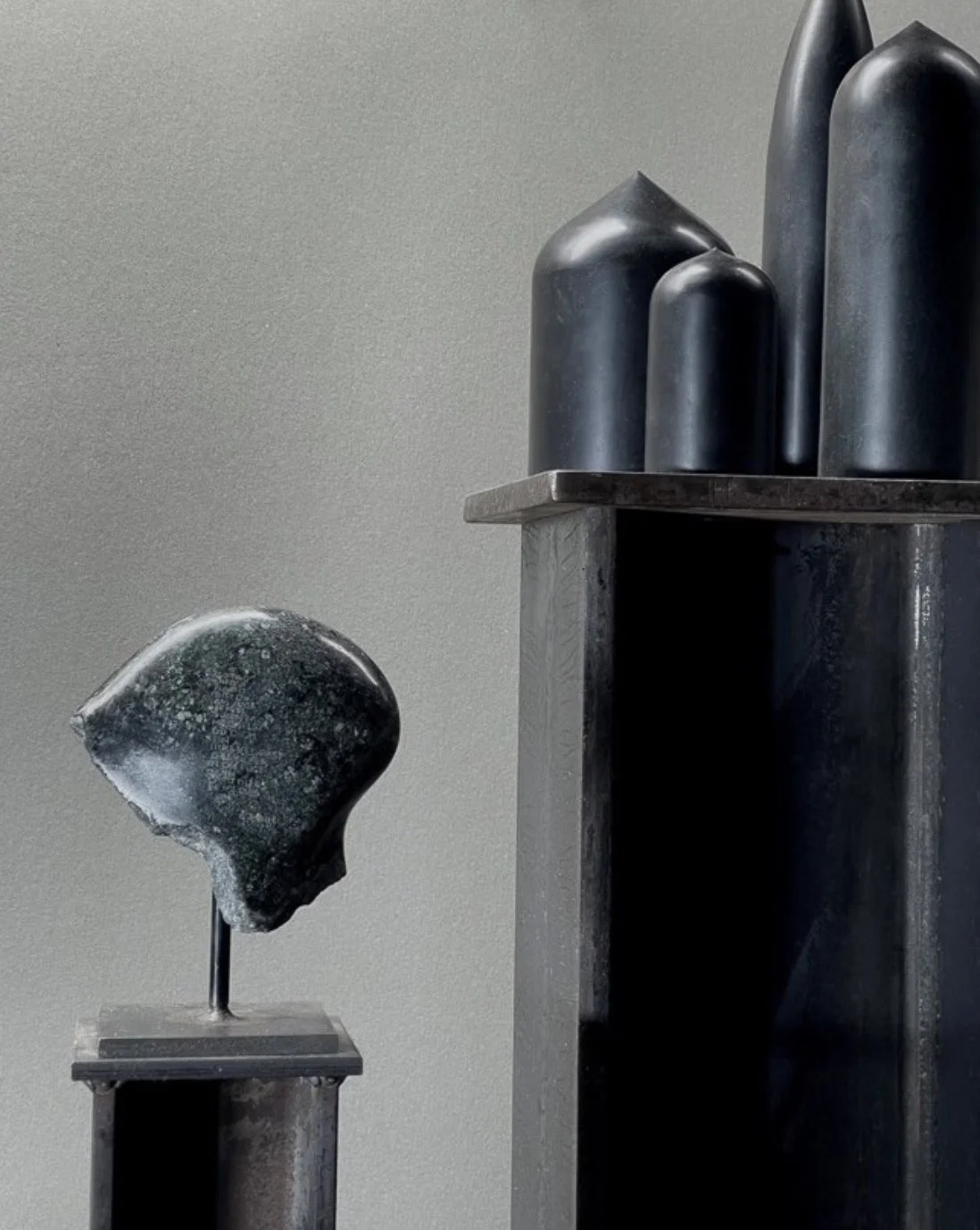 Dark, abstract sculptures resembling stones or pebbles on modern pedestals, displayed against a gray wall.