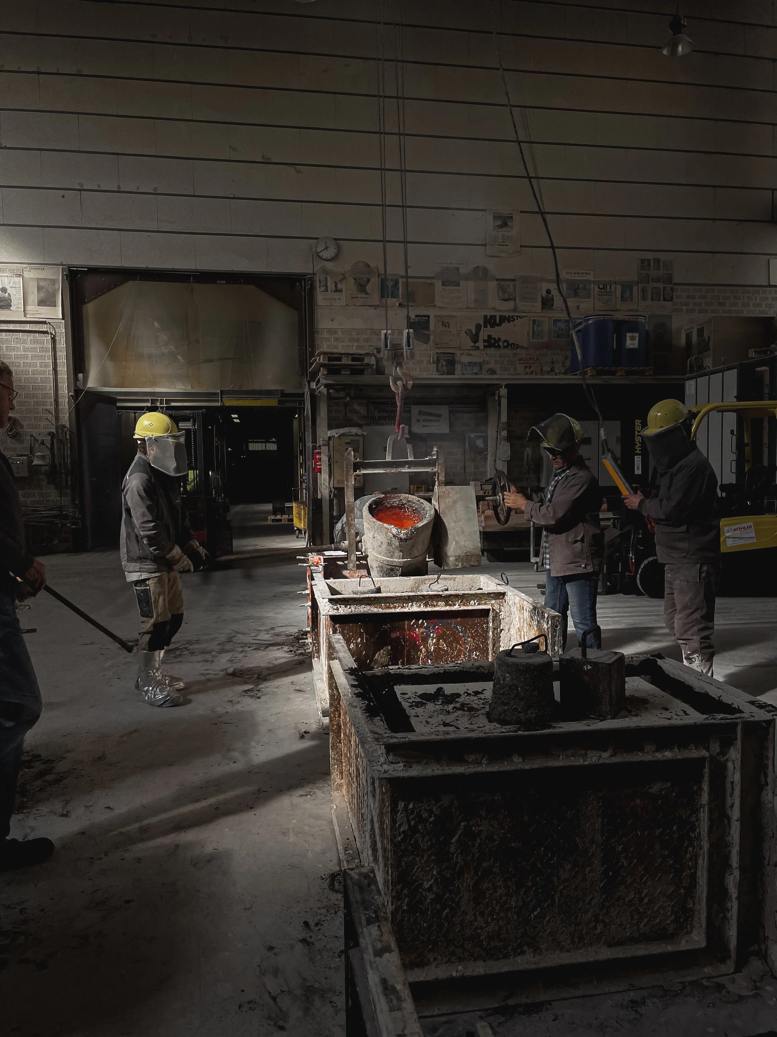Bronze cast workshop with workers wearing safety helmets and masks, working with molten bronze and equipment in an industrial setting.