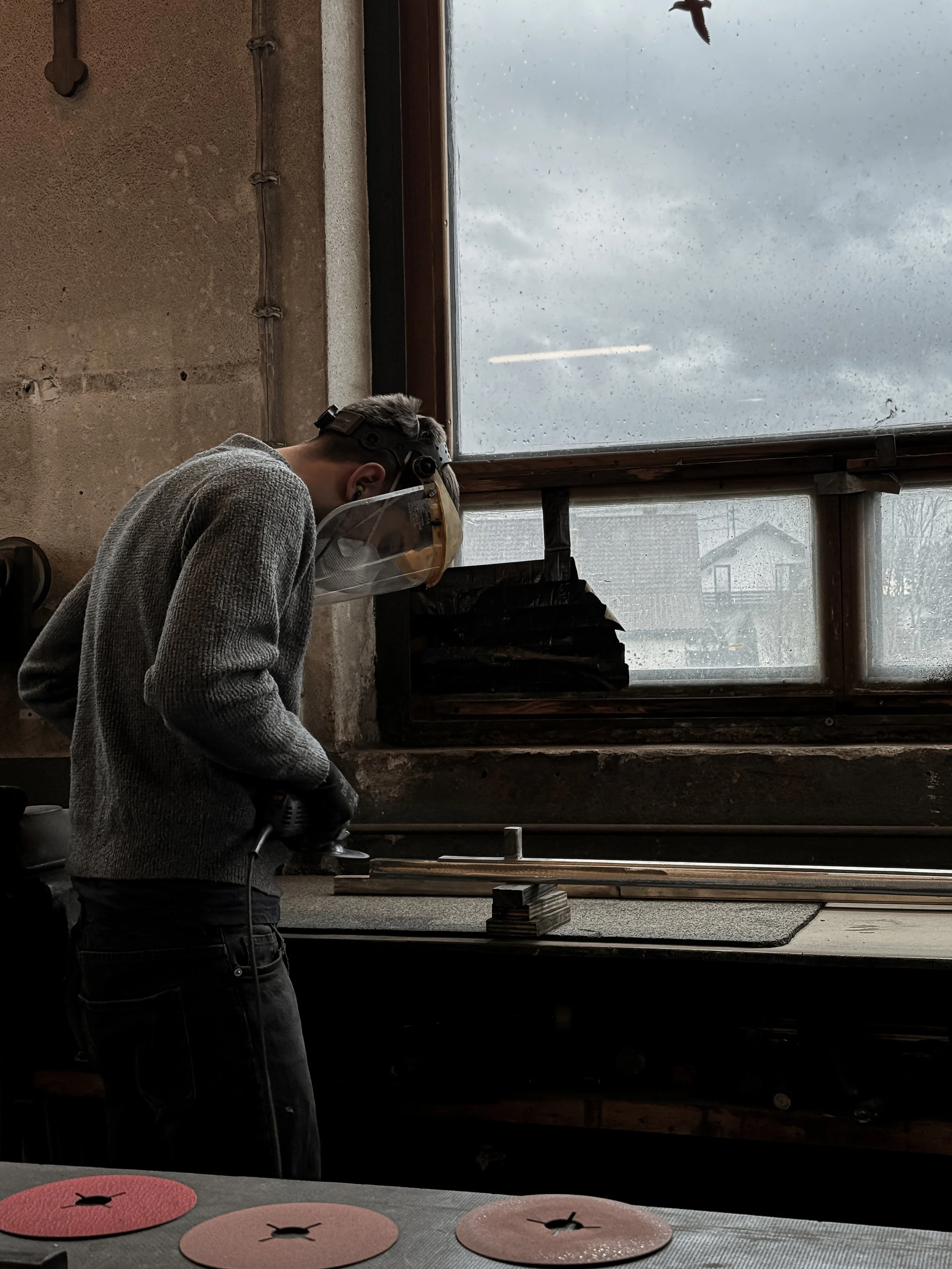 A person wearing a face shield, mask, and gloves working with a power tool at a workbench in a workshop, with a window showing a rain-drenched outside scene.