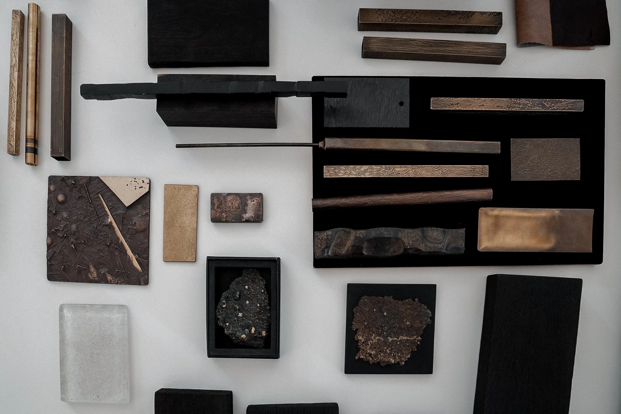 Collection of art samples, including textured stones, wood pieces, metal bars, and plaques, arranged on a flat surface.