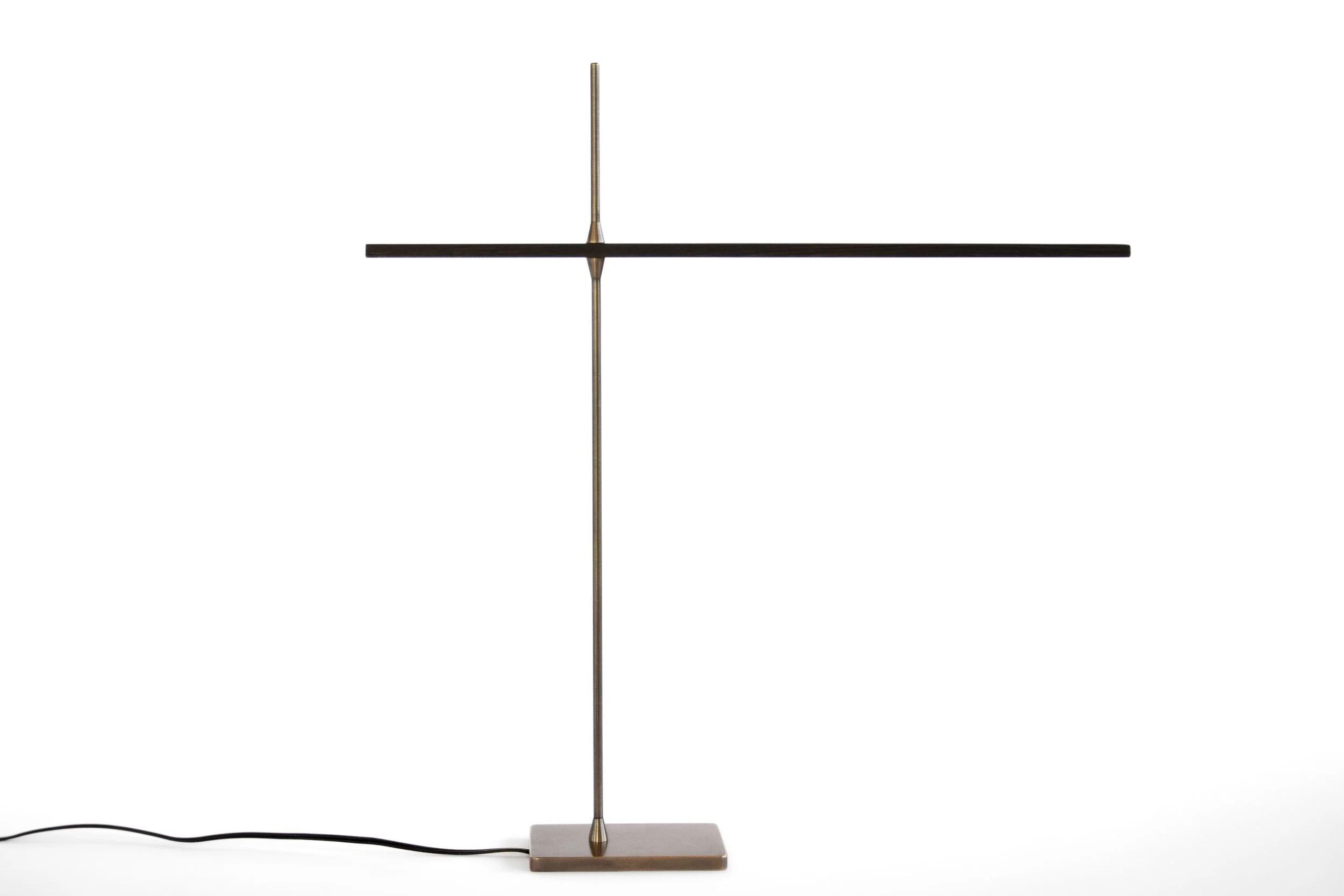 A modern, minimalist desk lamp with a vertical metallic pole, a horizontal wooden arm, a square brass base, and a black cord extending from the base.