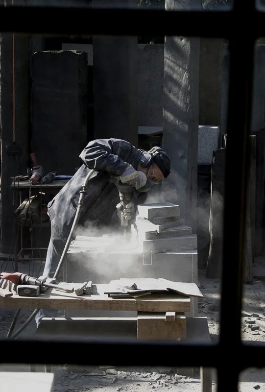A worker wearing protective gear is grinding a stone sculpture, creating a cloud of dust in a sculpture workshop.