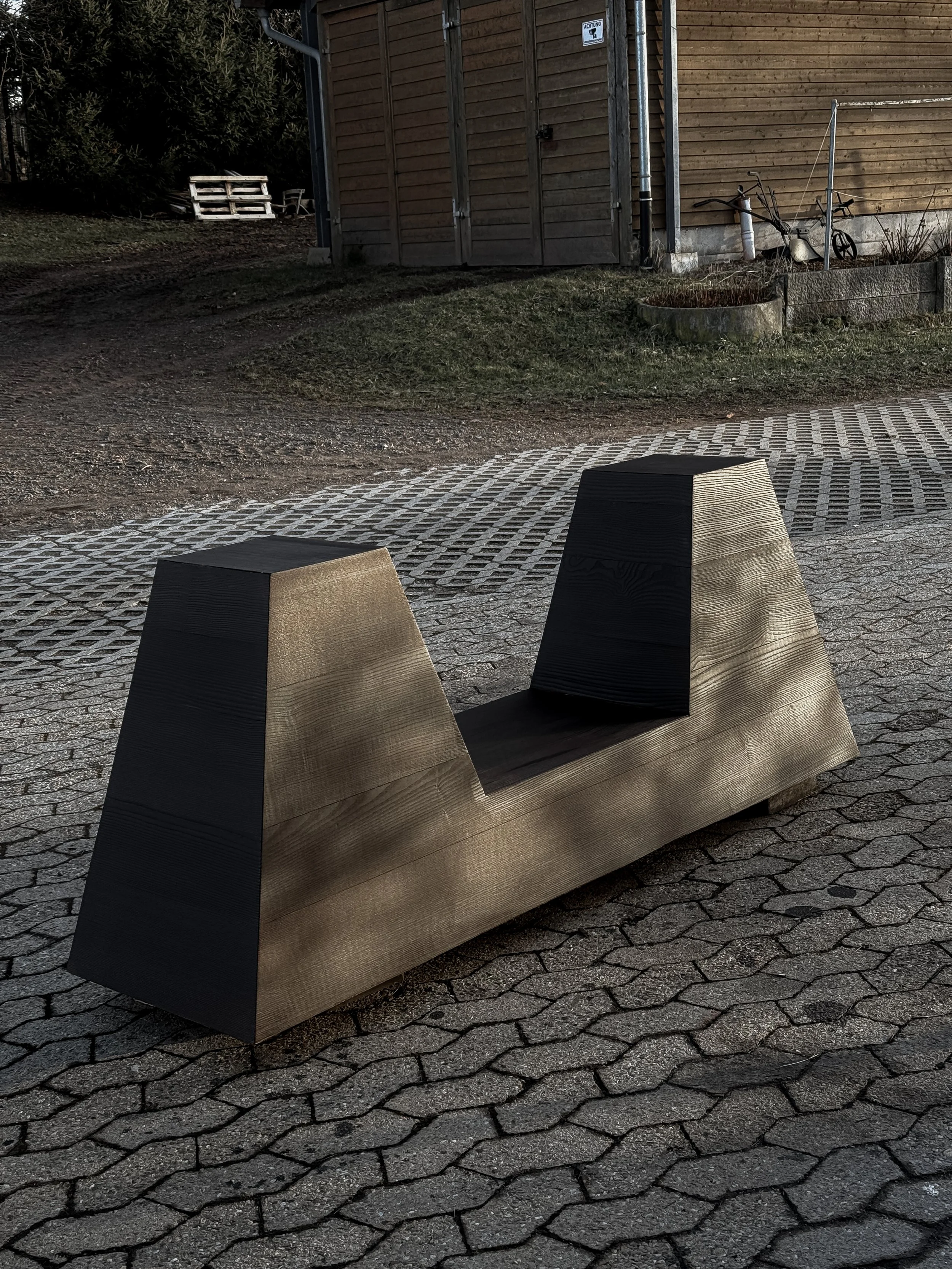 A modern, minimalist wooden table base with a geometric design located outdoors on a paved surface.