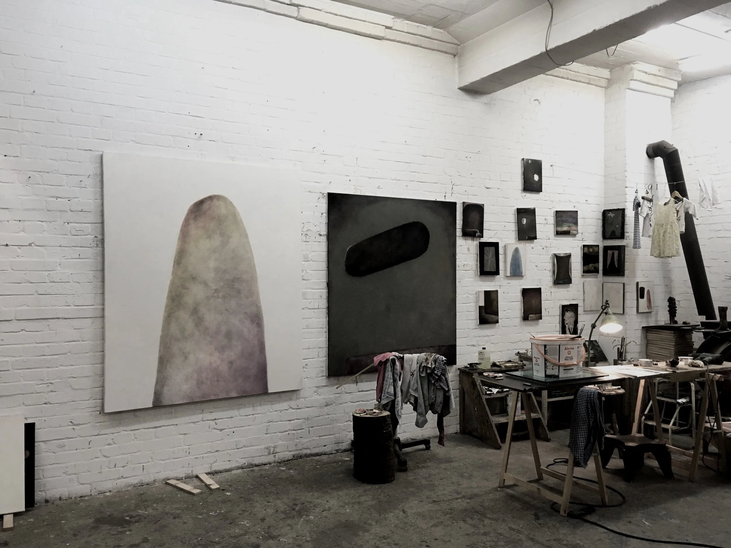 An art studio with white brick walls displaying various paintings and artworks, a work table with art supplies, and a black chimney pipe with clothes hanging.