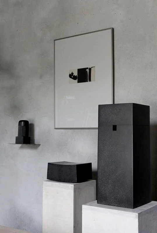 Minimalist interior with a framed abstract art piece, a small black  sculpture on a shelf, and three black modular geometric sculptures on white pedestals against a concrete wall.