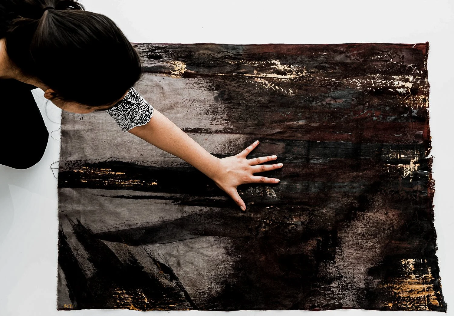 A person with dark hair and a patterned sleeve stretches out their arm on a large, abstract painting with dark colors and textured brushstrokes.