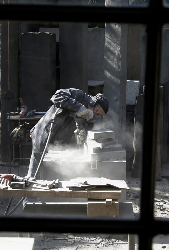 A worker wearing safety gear, including gloves and goggles, is using a tool to cut or shape a stone or concrete block at a construction or sculpture site, with dust and debris around.