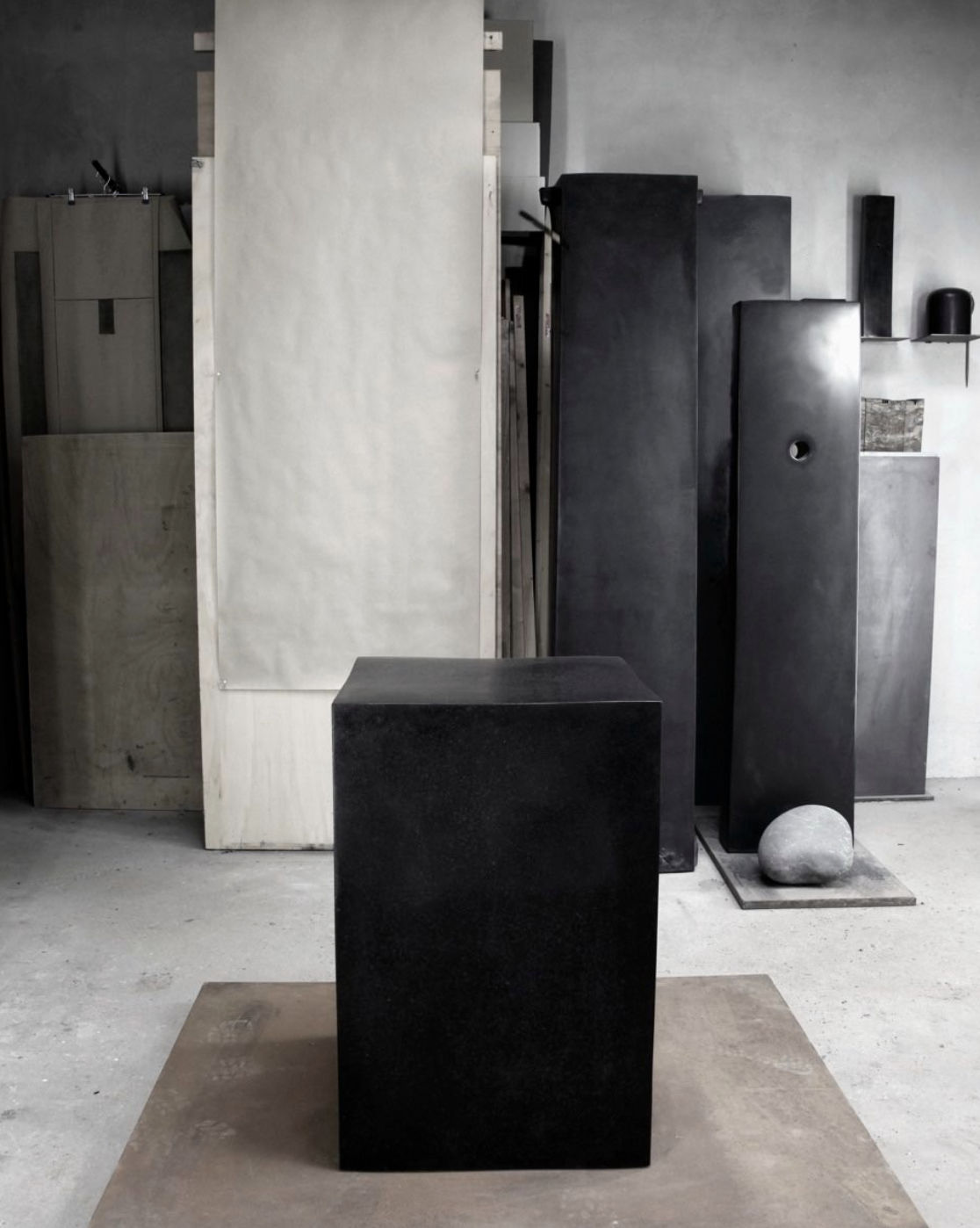 Art studio with minimalist sculptures, including a black rectangular block in the foreground and various tall black and light wood sculptures and objects against a gray wall in the background.