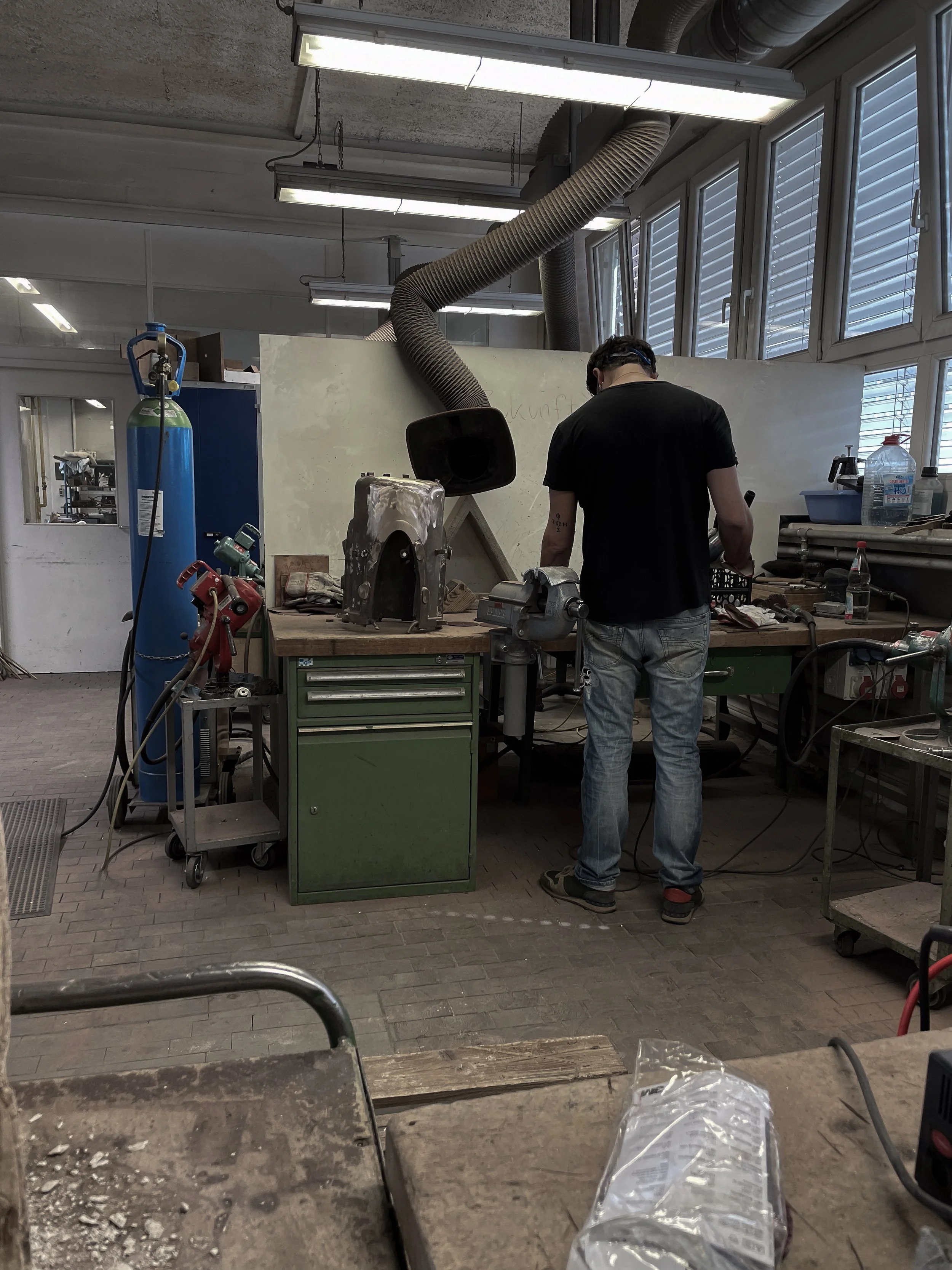 A person working at a cluttered workshop bench with tools, a welding helmet, and a large vent hose, near a window with blinds.