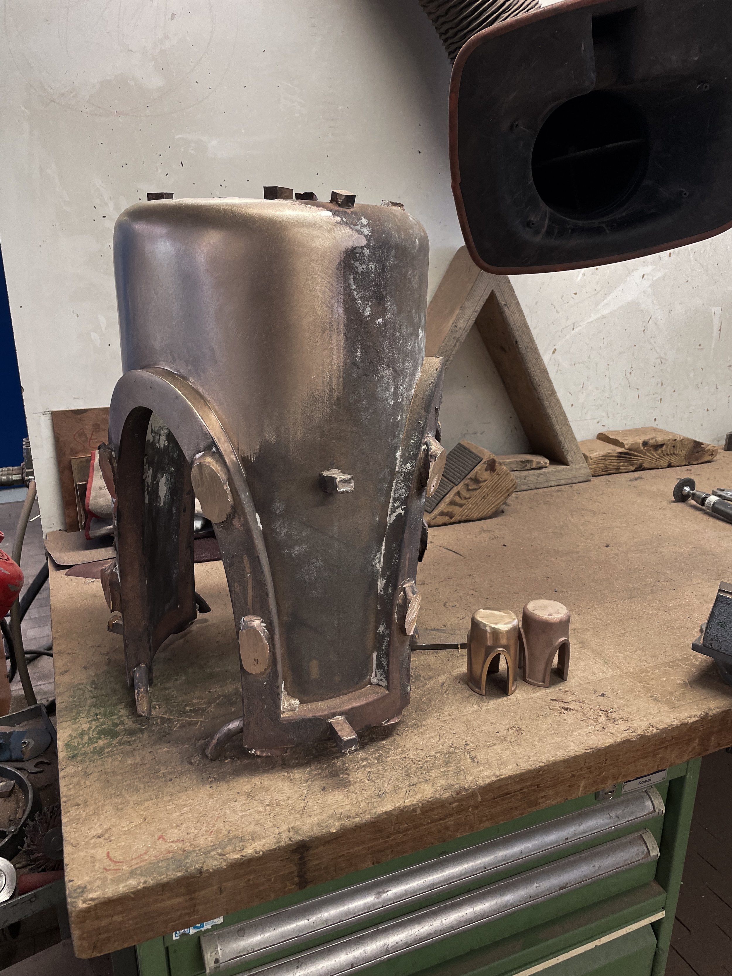 Metal sidetable on a wooden workbench, with two small bronze colored stools or components nearby.