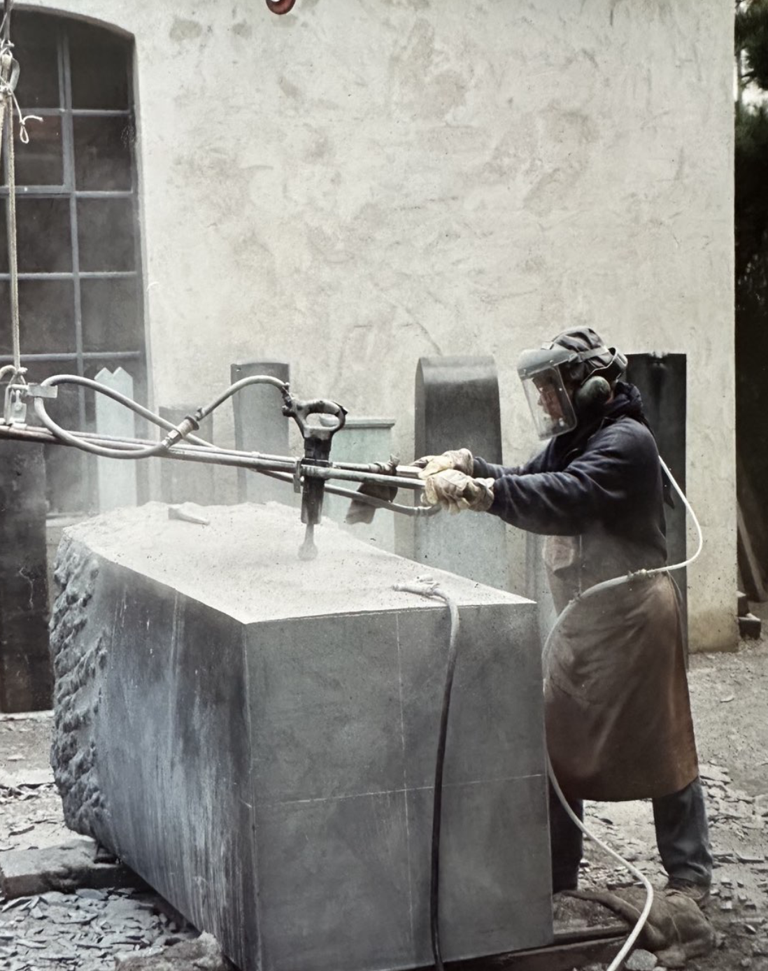 A person wearing a protective helmet, face shield, gloves, and apron is operating a large power tool, possibly a stone or concrete saw, cutting through a stone block outdoors.
