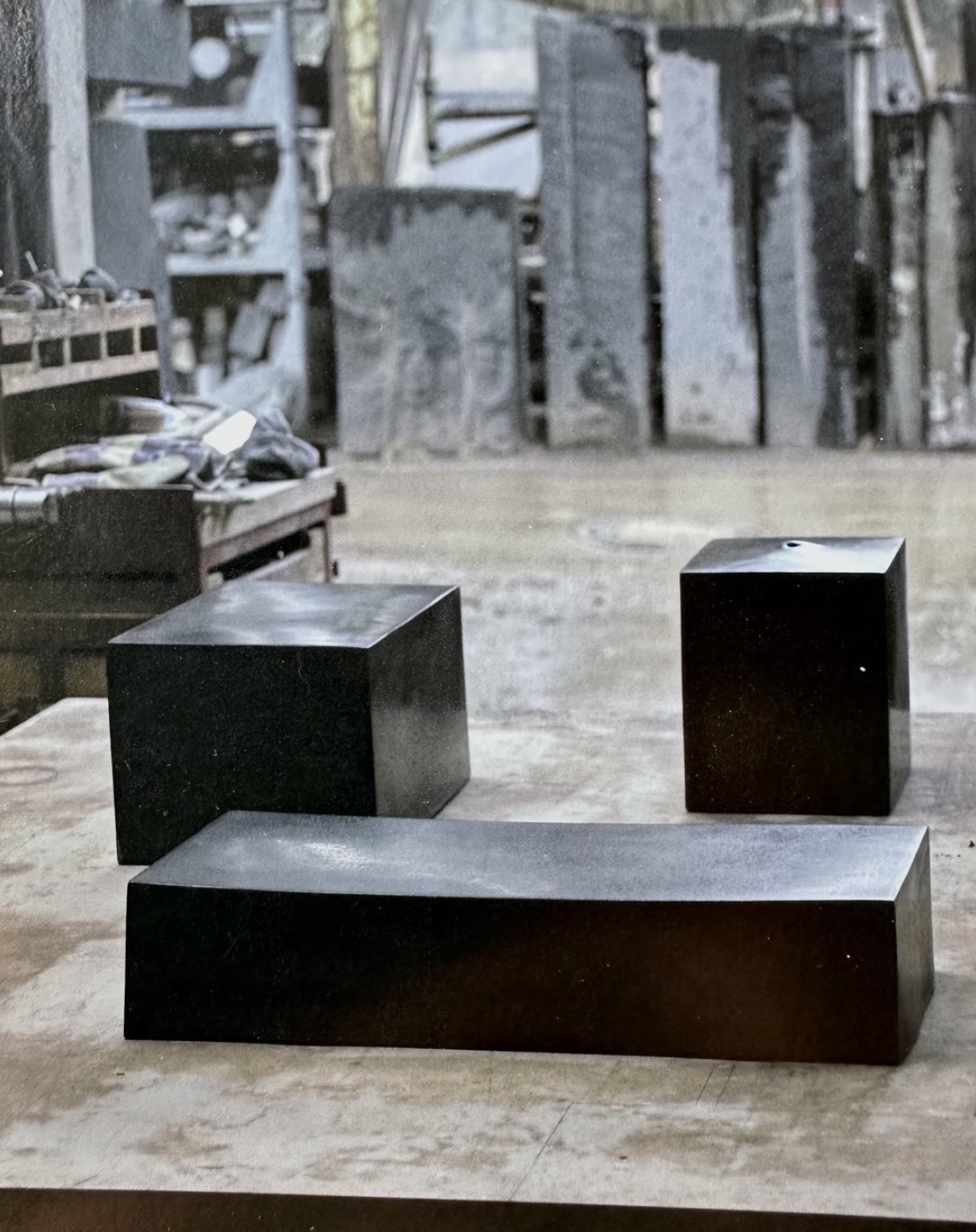 Three black rectangular and square objects arranged on a wooden surface, with an industrial workshop in the background.