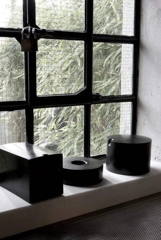 Three black geometric sculptures on a white ledge in front of a window with metal grid bars and green foliage outside.