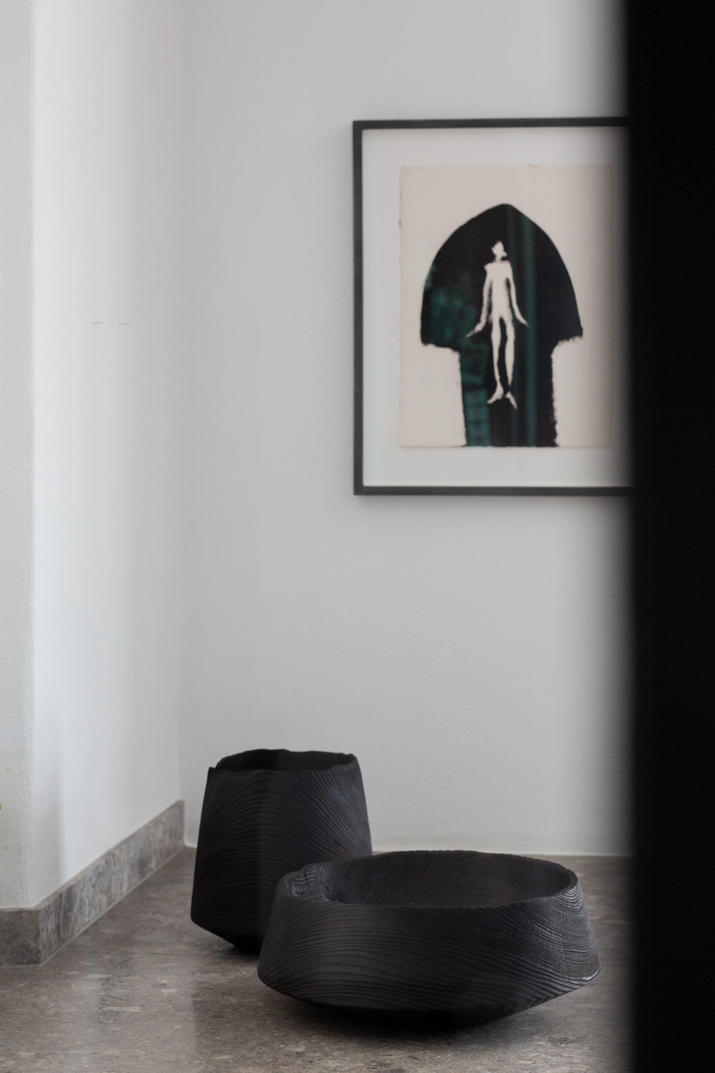 Minimalist interior with a framed abstract artwork of a human figure and rectangular shapes on a white wall, and two black textured pottery bowls on a gray floor.