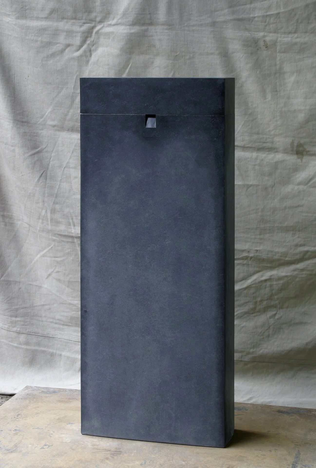 A tall, rectangular, dark stone  structure with a small square opening near the top, placed on a wooden surface with a crumpled beige fabric backdrop.