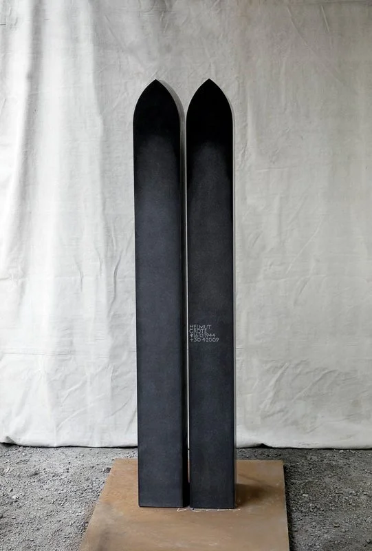 A pair of black skis standing upright on a wooden base against a plain, light-colored fabric backdrop.