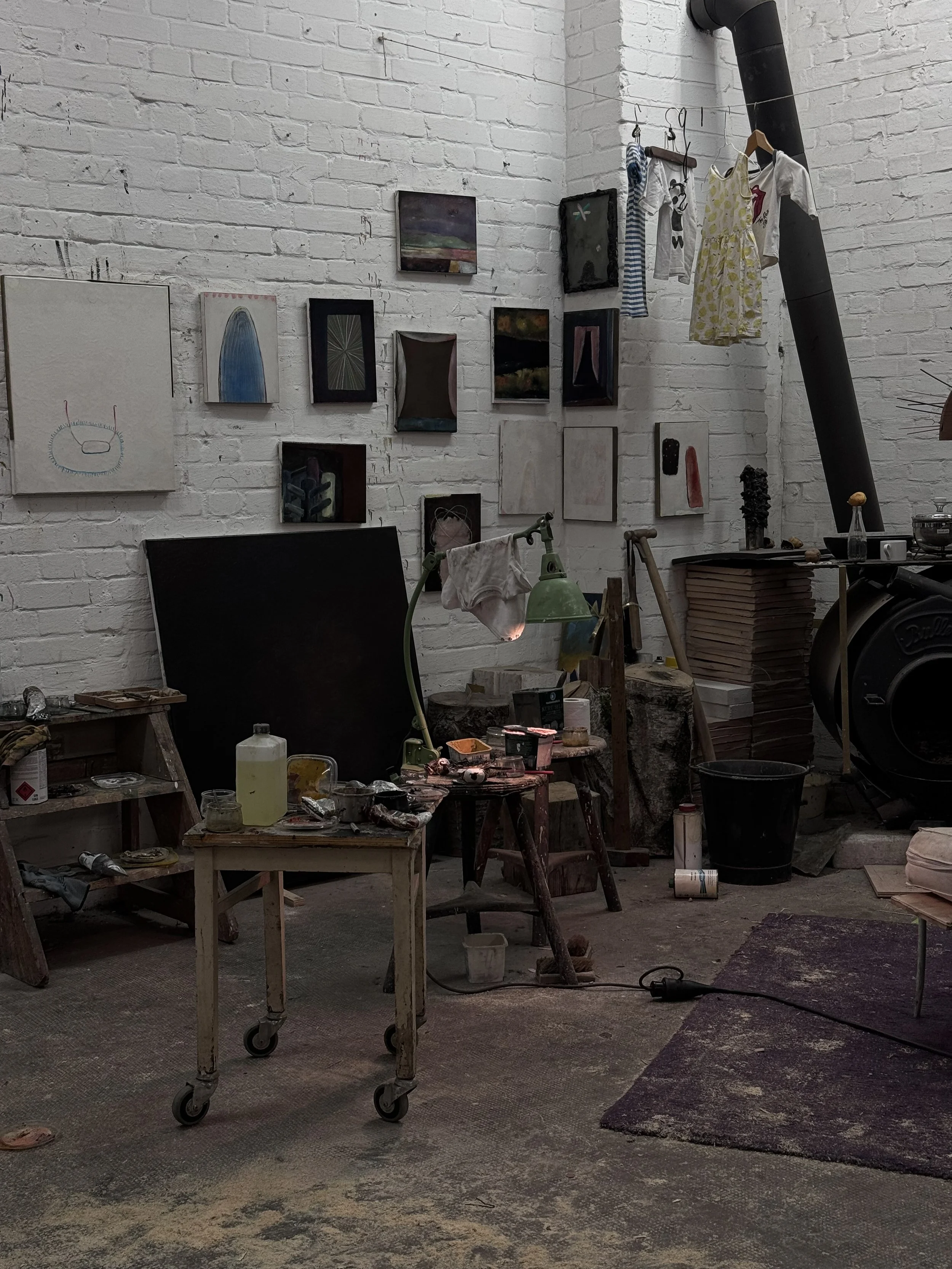 An artist's studio with a white brick wall featuring multiple small paintings and art pieces. The space includes tables with art supplies, a green desk lamp, and clothes hanging from a pipe. The floor is dusted with paint or dirt, and there is a wood stove in the corner.