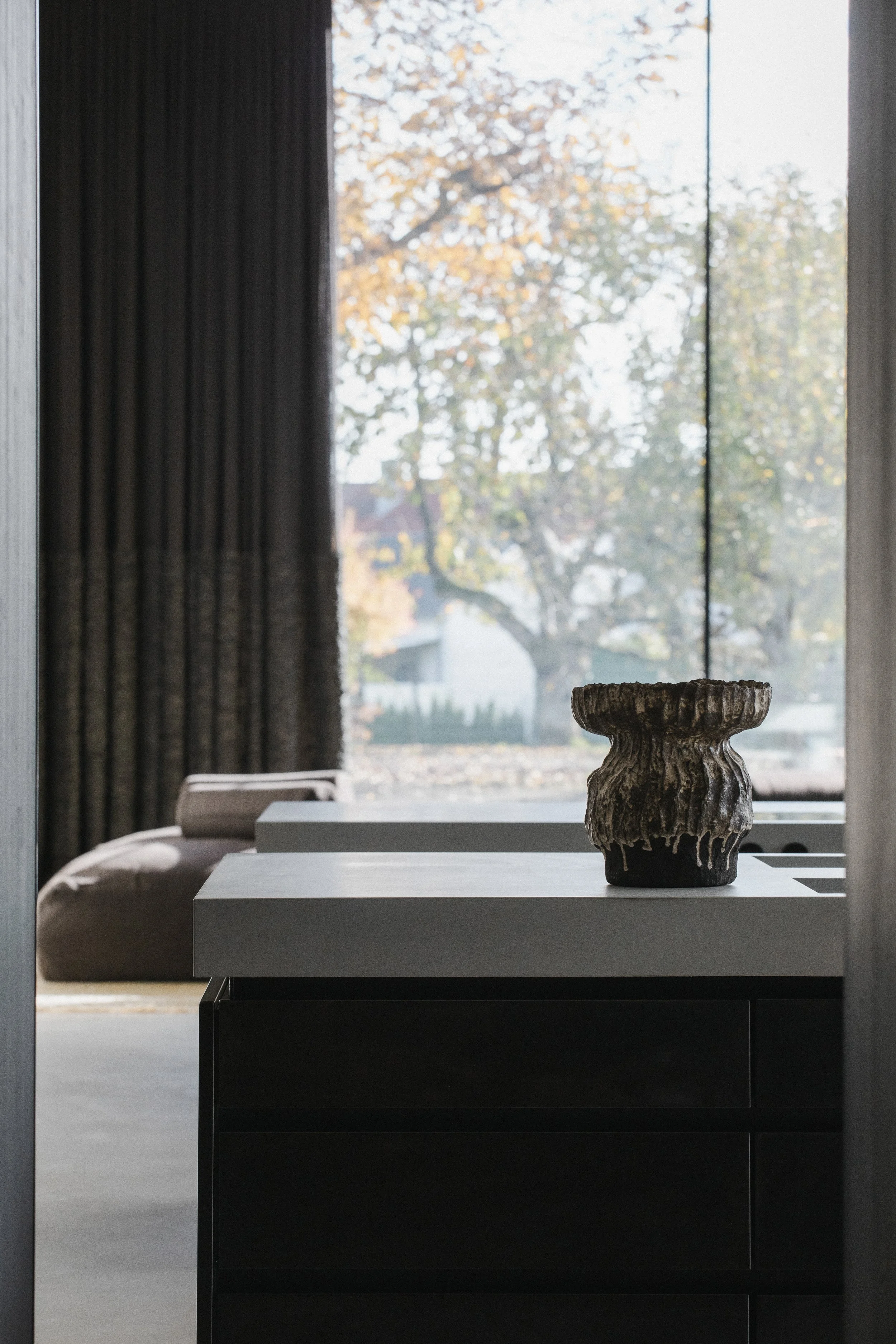 A ceramic vase on a white countertop inside a modern room, with large windows showing autumn trees outside.