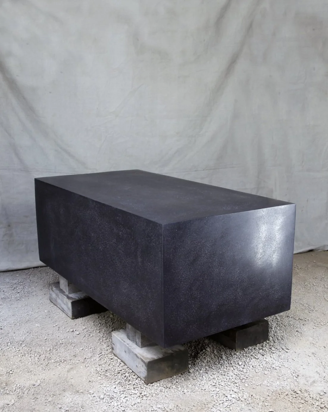 A large black rectangular stone block supported by concrete and wood blocks, placed on a gravel floor against a plain fabric backdrop.