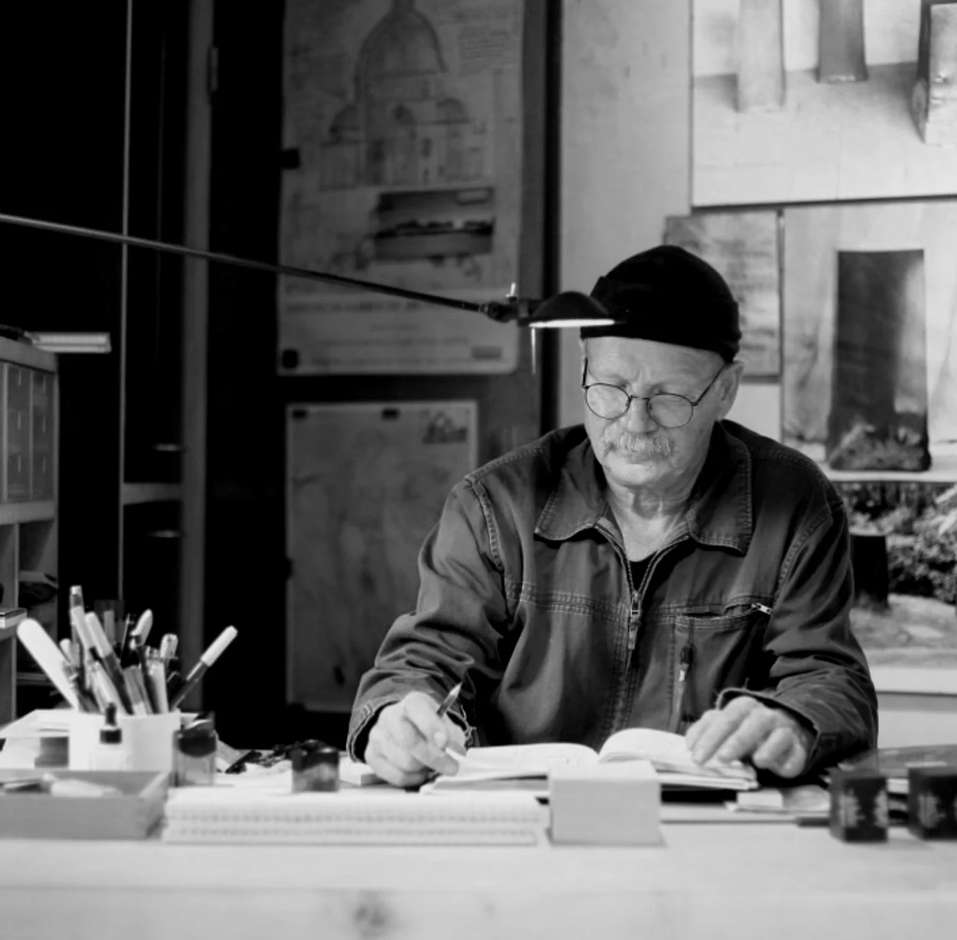 An elderly man with glasses, a mustache, and a black hat, wearing a casual jacket, sitting at a desk writing in a notebook with various art supplies nearby, in a room with maps and posters on the wall.