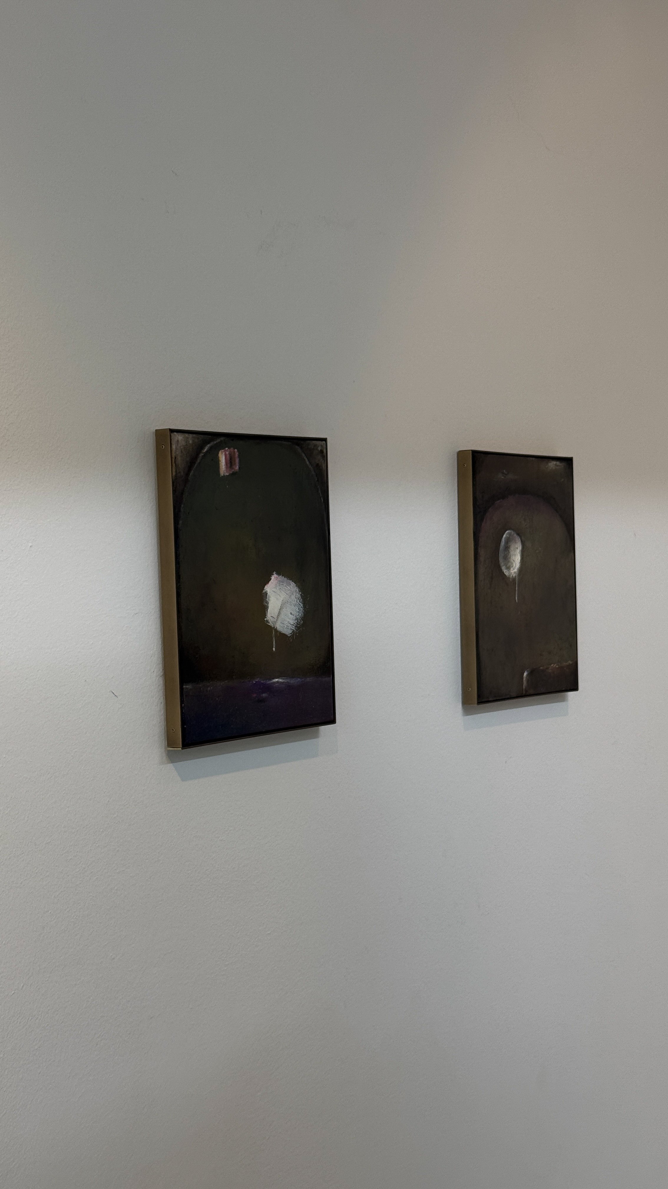 Two abstract paintings hung on a plain white wall. The paintings have dark, muted tones with some white and pale colors, and are framed in metallic frames.