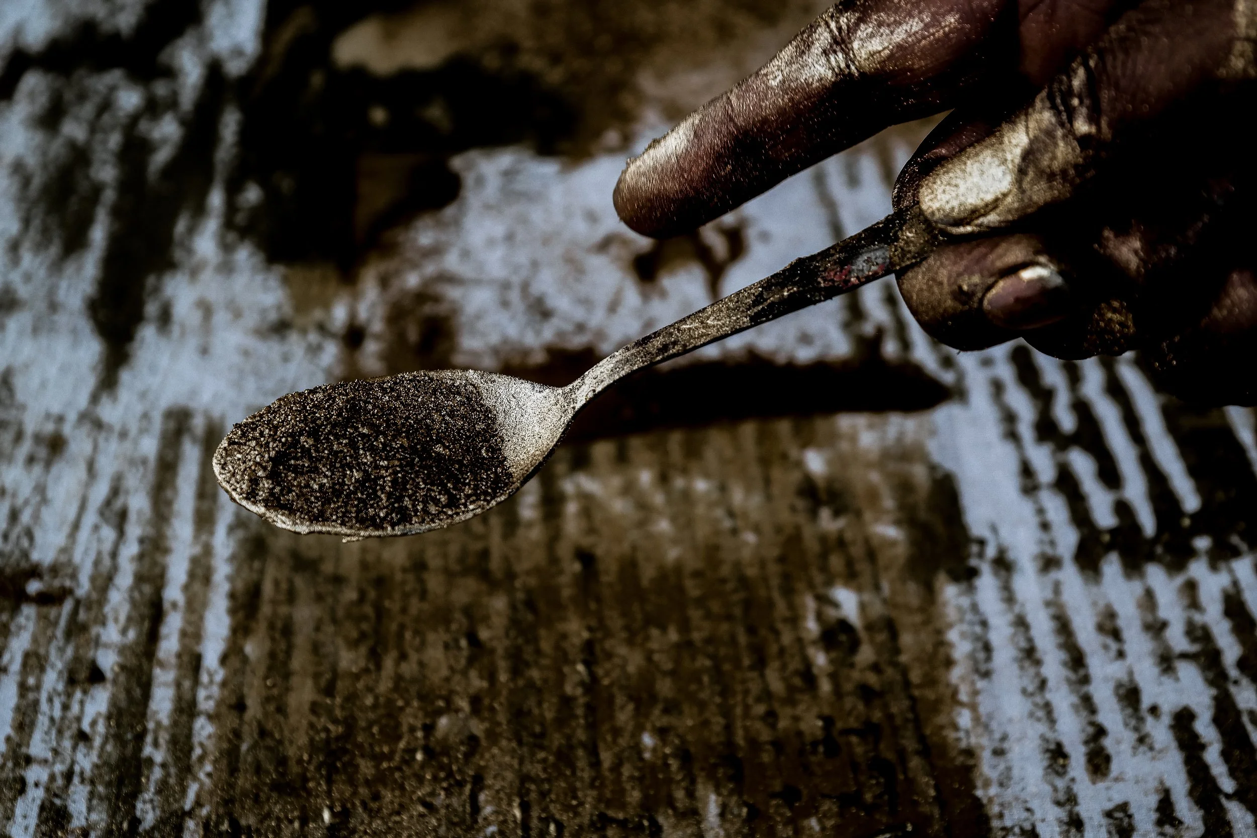 A close-up of a hand holding a small spoon filled with pigments, with a rustic  surface in the background.