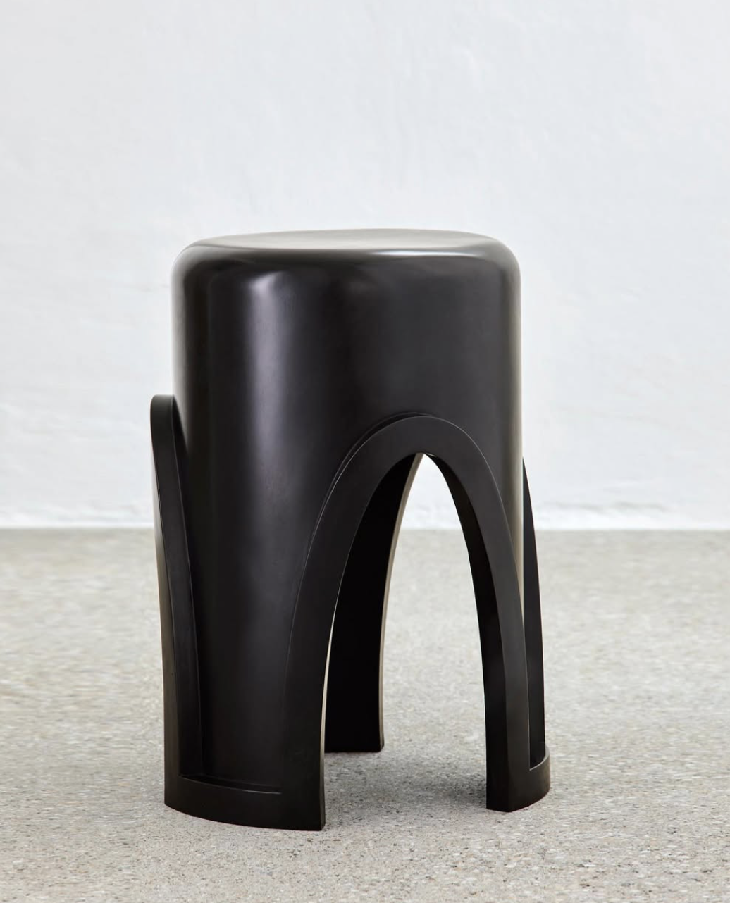 A black modern stool with a rounded top and arched supports on a light-colored floor against a white wall.