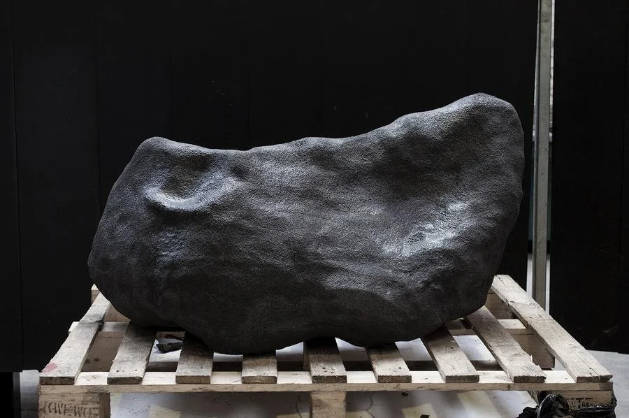 Large, smooth, black ceramics rock resting on a wooden pallet against a black background.