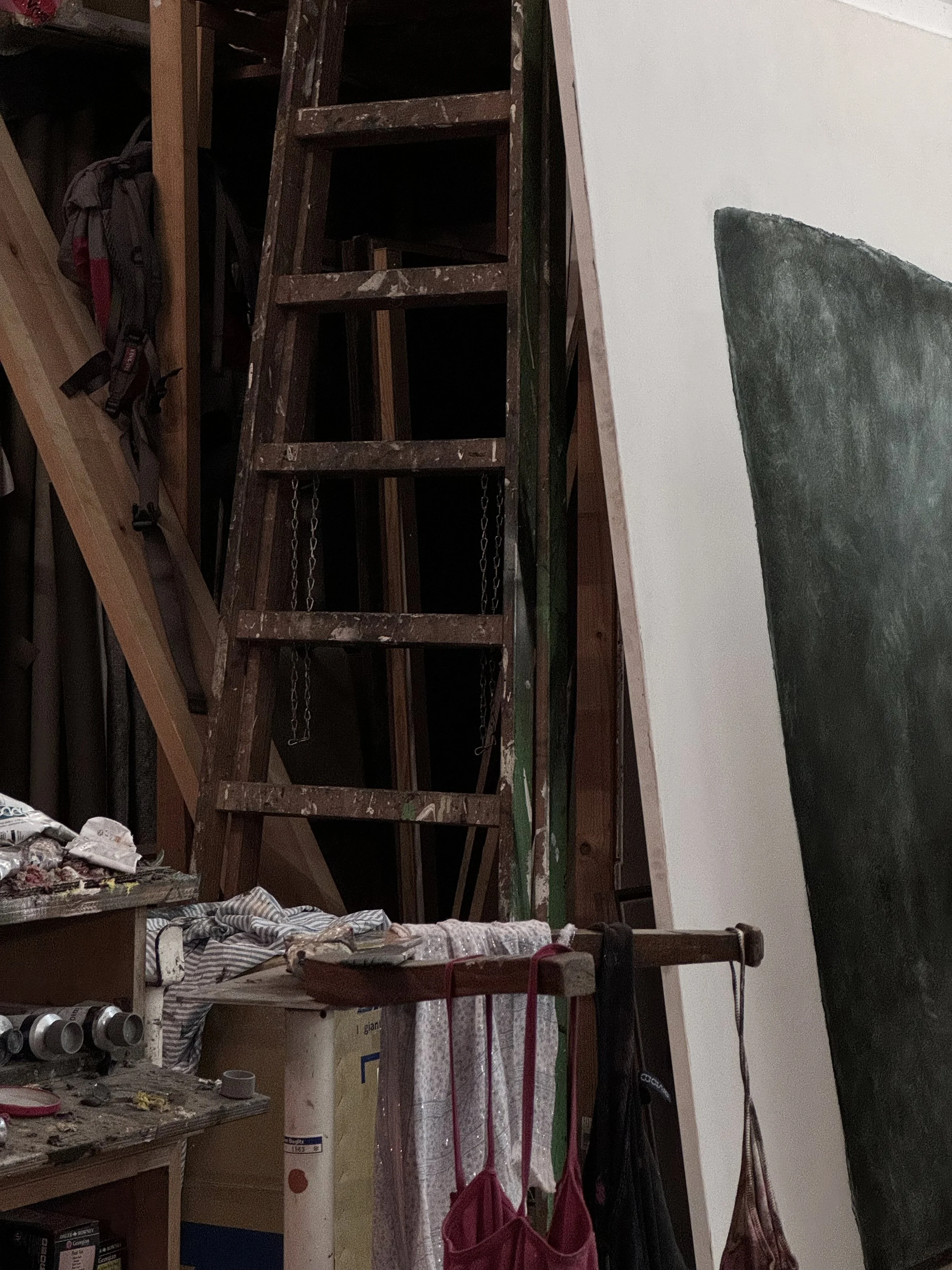 A wooden ladder leaning against a wall in an art studio with supplies and hanging bags in the foreground.