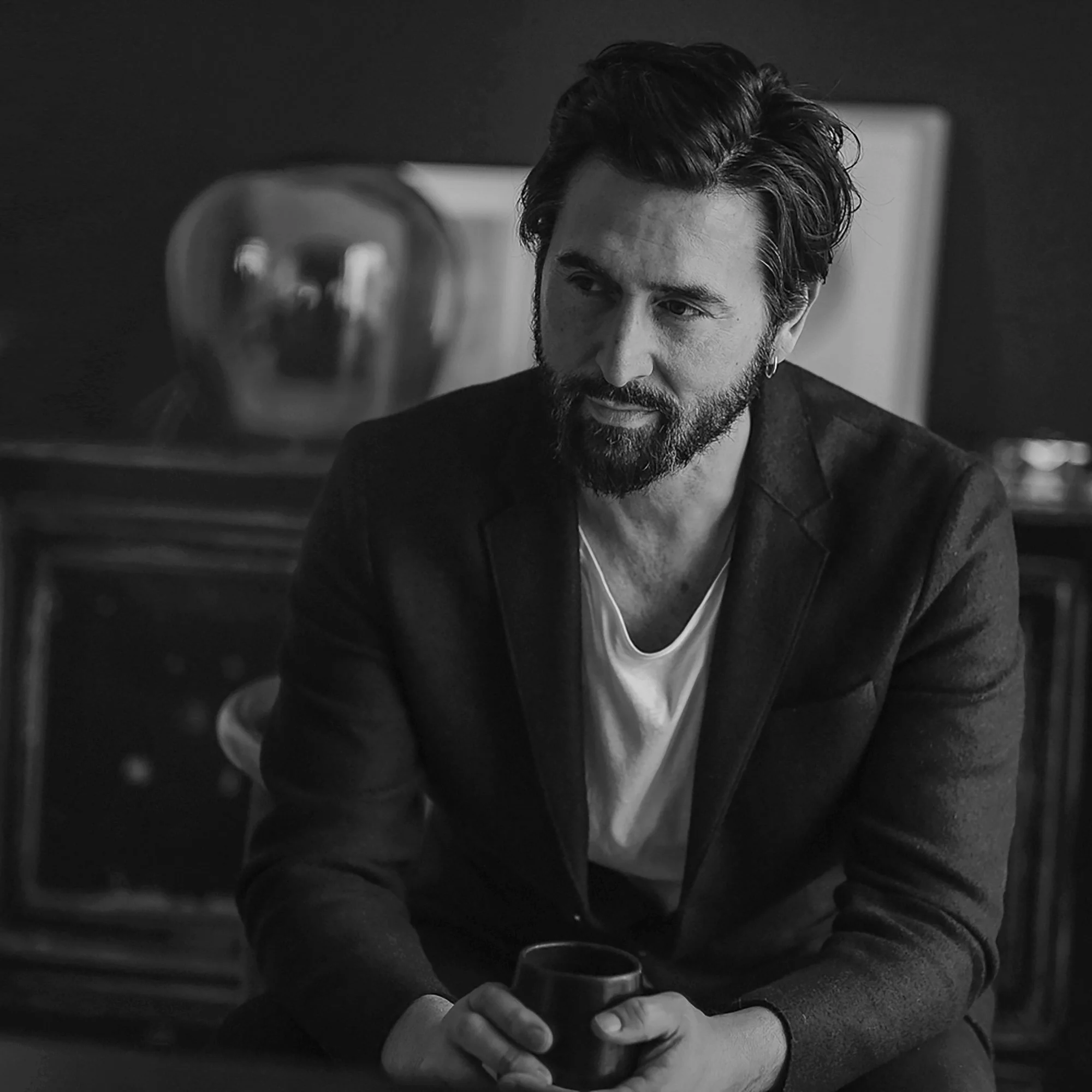 A man with a beard and wavy hair, wearing a dark blazer and a white shirt, sitting indoors holding a glass cup.