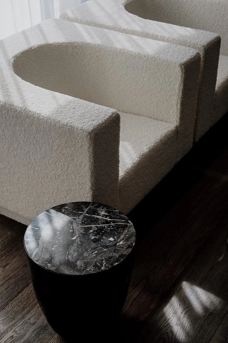 A modern interior scene with a textured cream-colored armchair labeled as a Wassily Chair, paired with a round black marble-top side table. The furniture is on dark hardwood flooring near a window with sunlight streaming in.