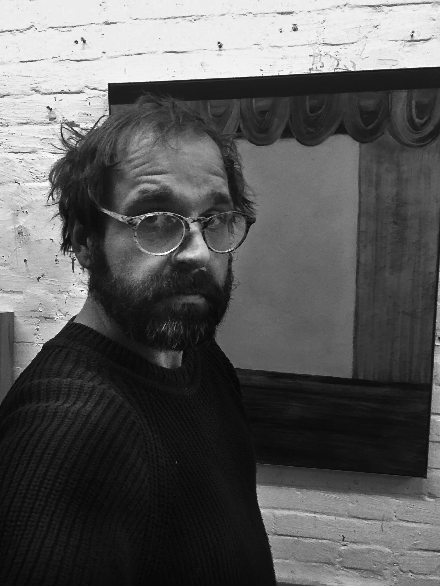 A man with glasses and a beard taking a selfie in front of a mirror in a room with white brick walls.