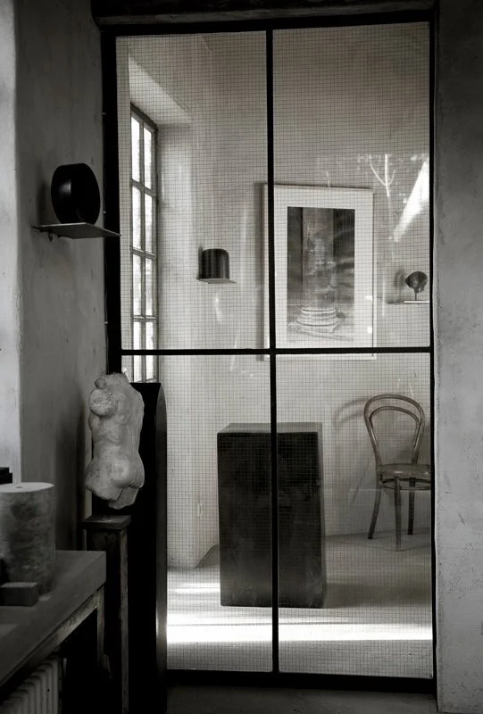 Interior view of a room, seen through a glass panel with black grid divisions, displaying a mannequin torso, a wooden chair, framed artwork, and various decorative objects.