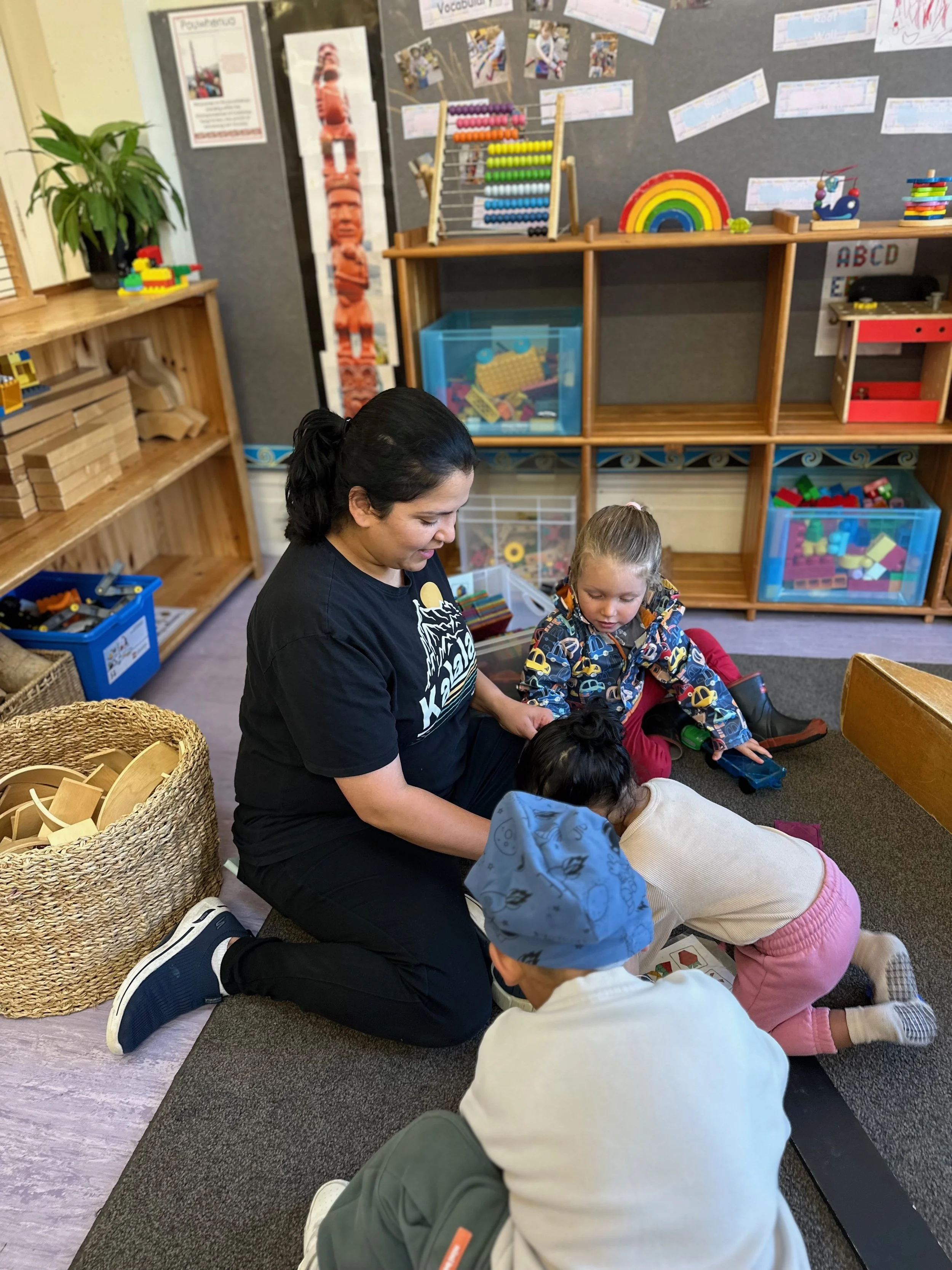 A teacher and four young children engaging in a play activity on the floor of a classroom, surrounded by shelves with toys and educational materials, with a colorful weather chart and books on the wall.
