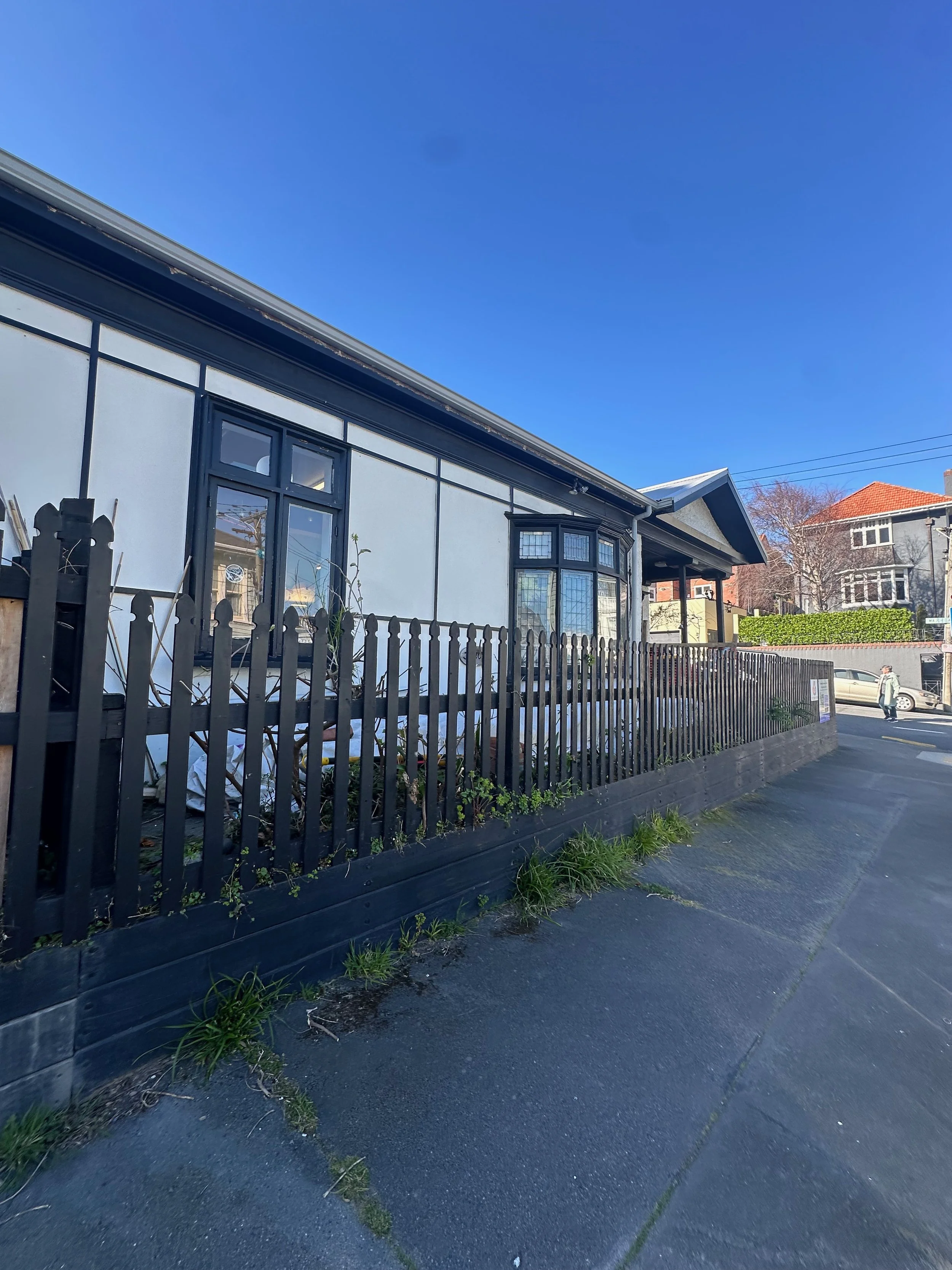 A street view of a building with a white exterior, black trim, and large window. There is a black wooden fence along the sidewalk with some small plants and weeds growing nearby. The sky is clear and blue, with neighboring houses visible in the backg