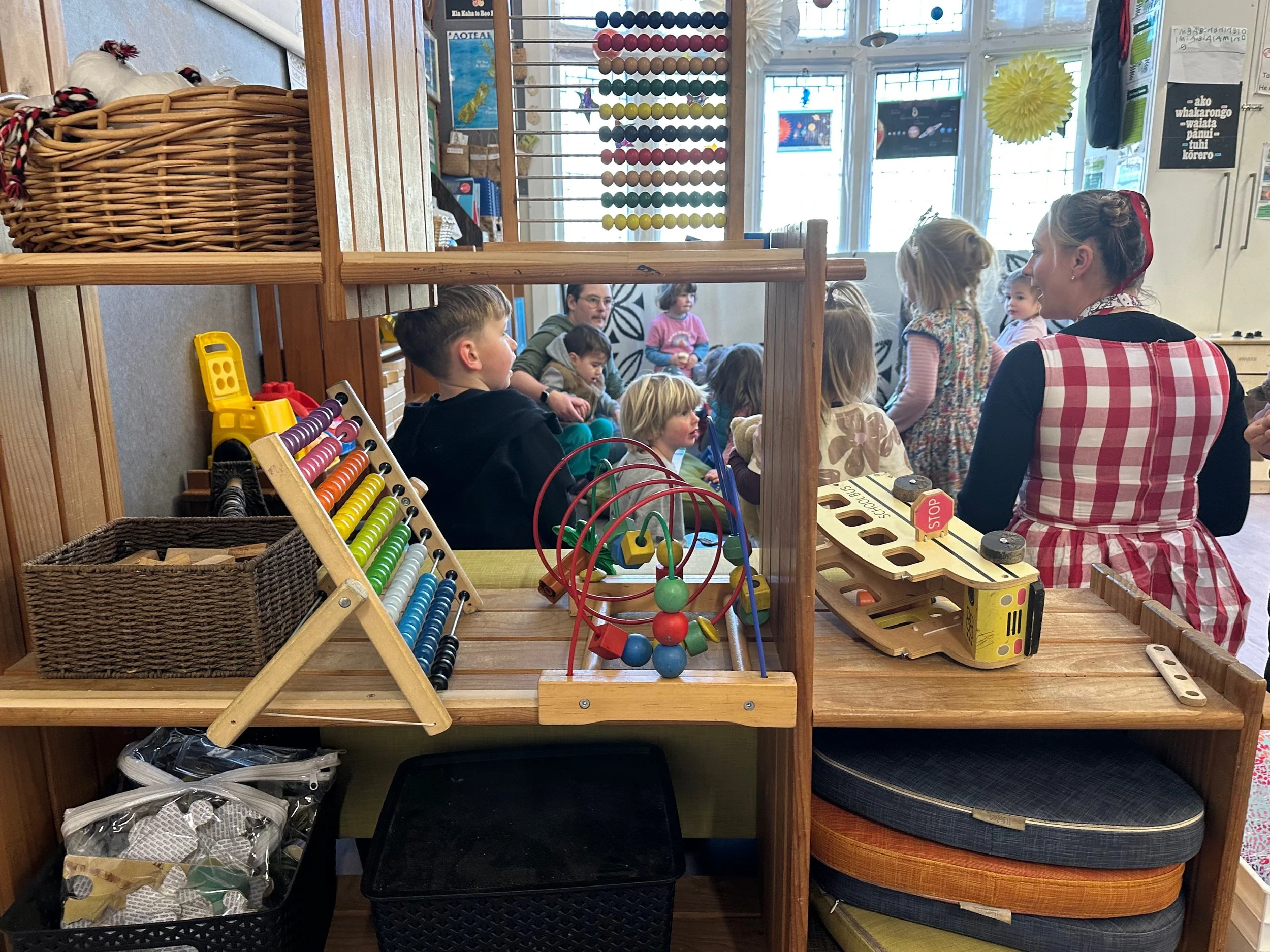 Children and adults gathered in a classroom or playroom, engaging in activities. Toys and educational materials are on shelves and tables, with a window in the background.