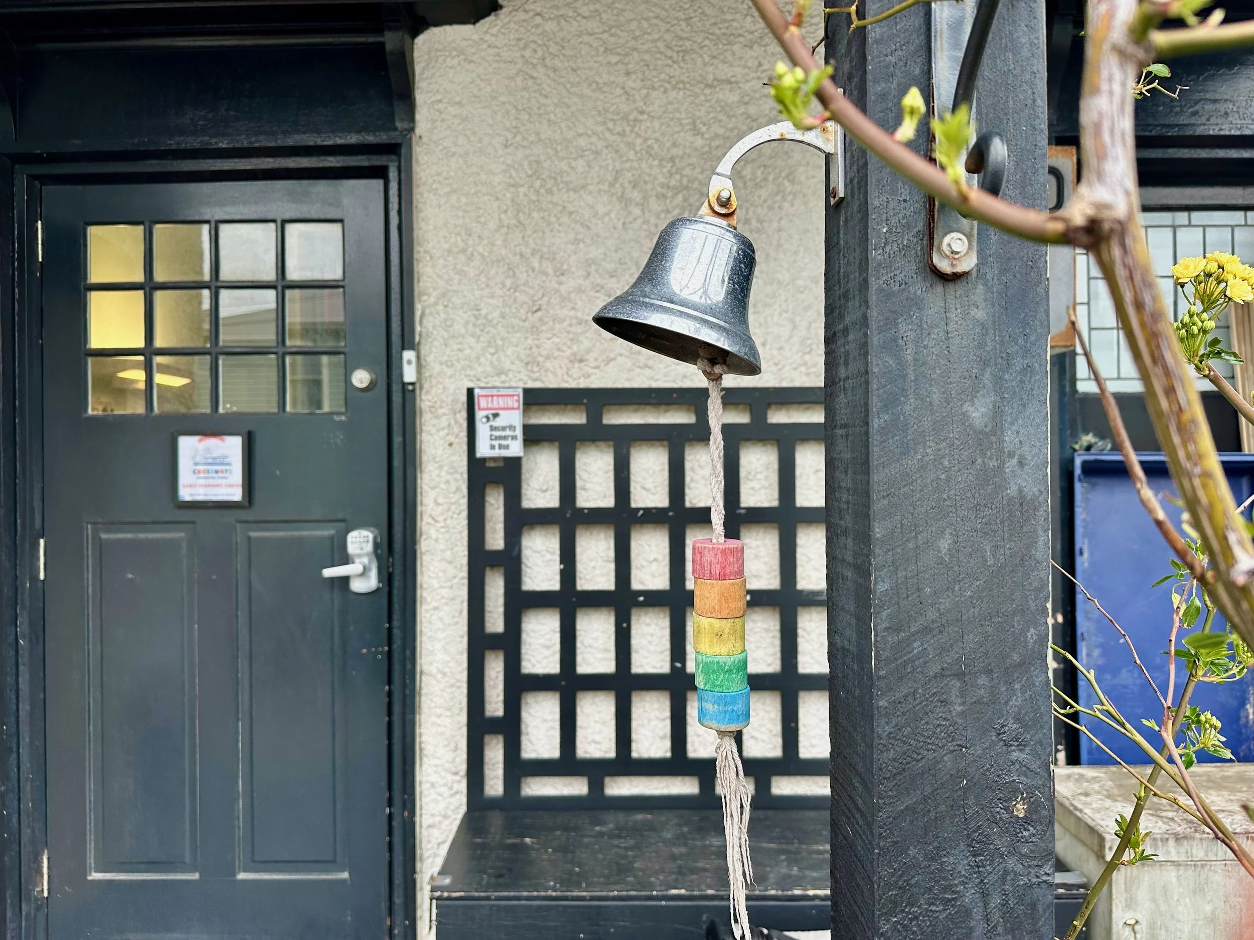 A black door with a window and a latch, a security warning sign, and a small bell hanging from a black post with colorful wooden beads on a rope. There are some yellow flowers and green leaves in the foreground.