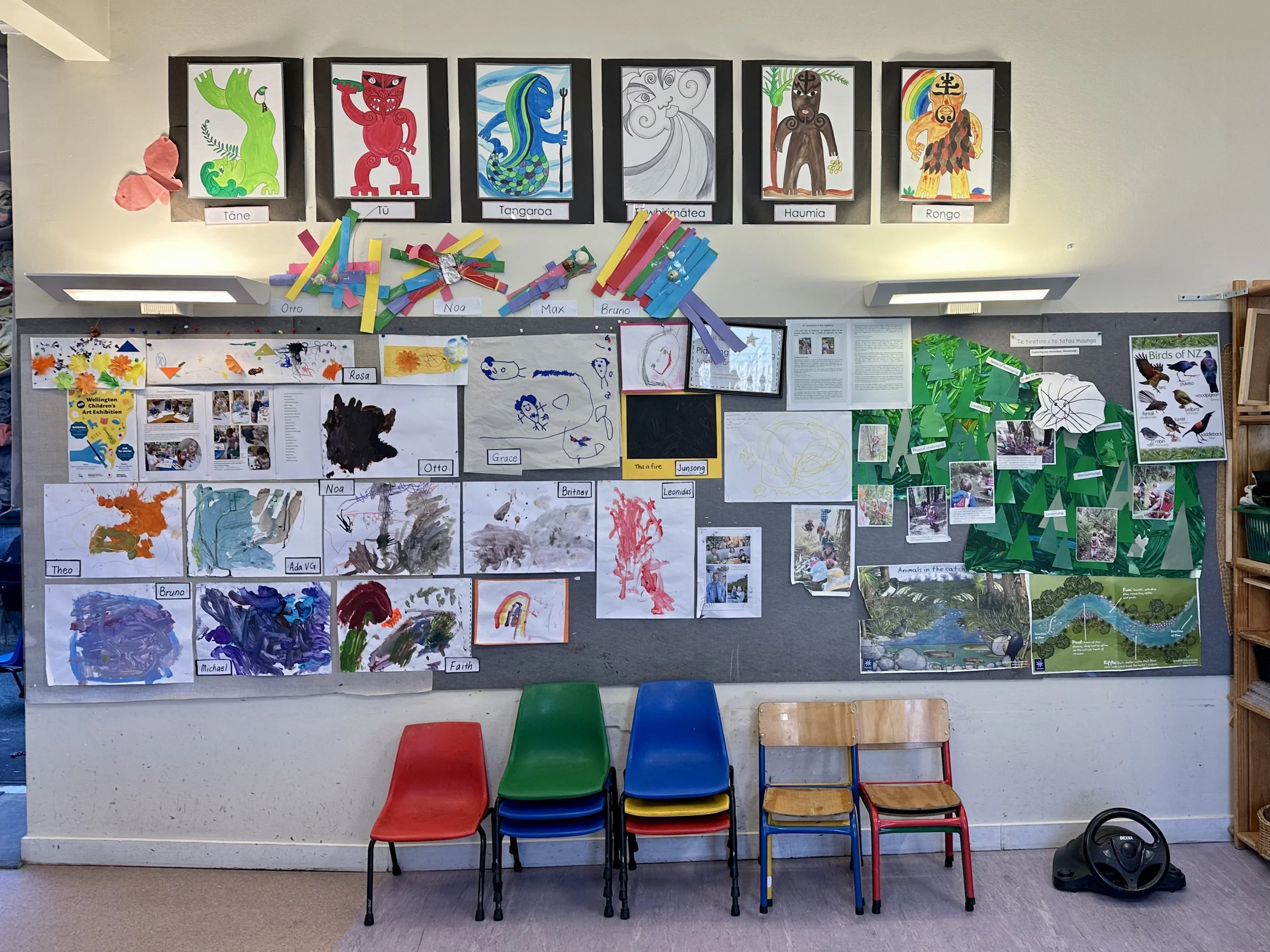 Classroom bulletin board with children's artwork, photos, and educational displays, featuring colorful drawings, paintings, and collages, alongside framed pictures of Maori gods, a bird poster, and nature scenes, with four small chairs in front.