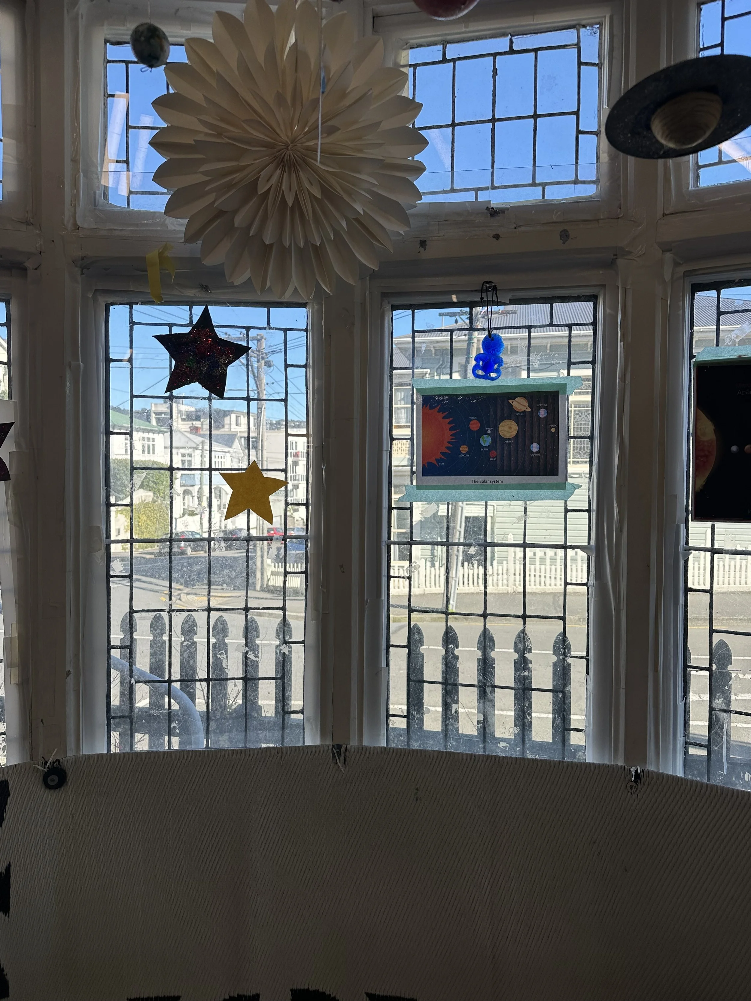 View of a classroom window with space-themed decorations, including stars, a large paper flower, and a solar system poster.