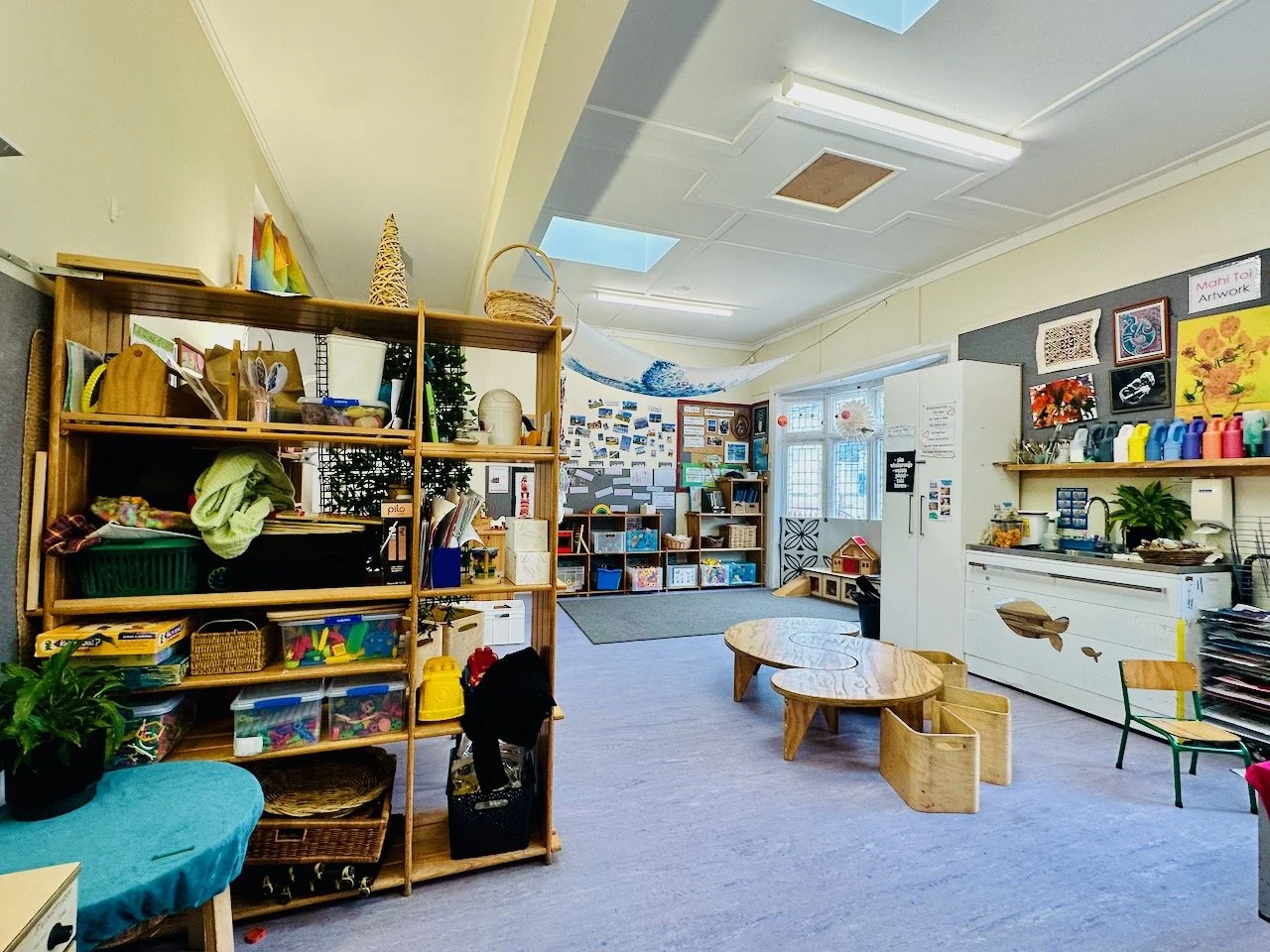 A colorful classroom with shelves filled with toys, books, and art supplies. There are small tables and chairs, a wall with art projects, and a window letting in natural light.